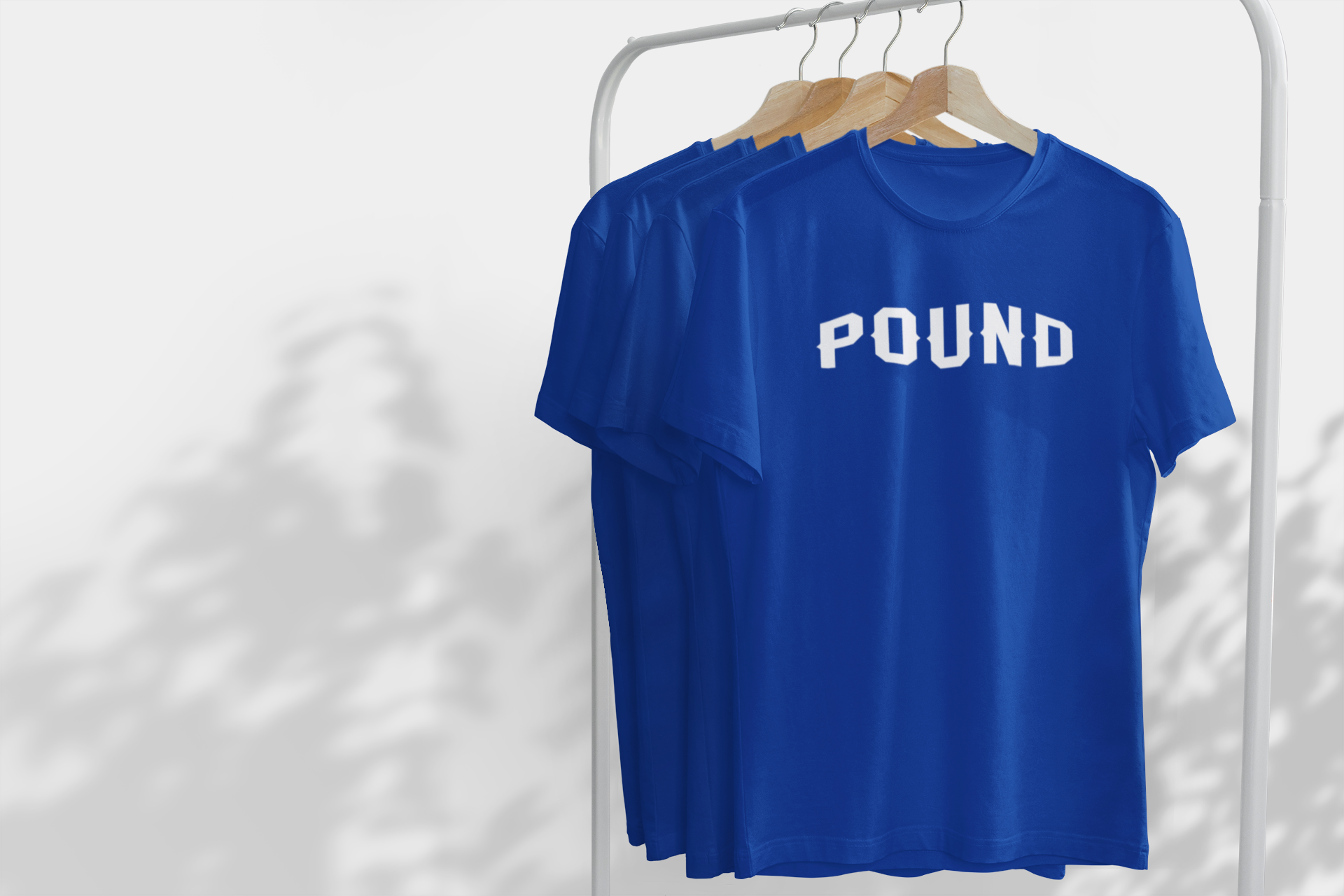 Three blue T-shirts hanging on a white clothing rack, with the front one displaying the word 'POUND' in white letters.