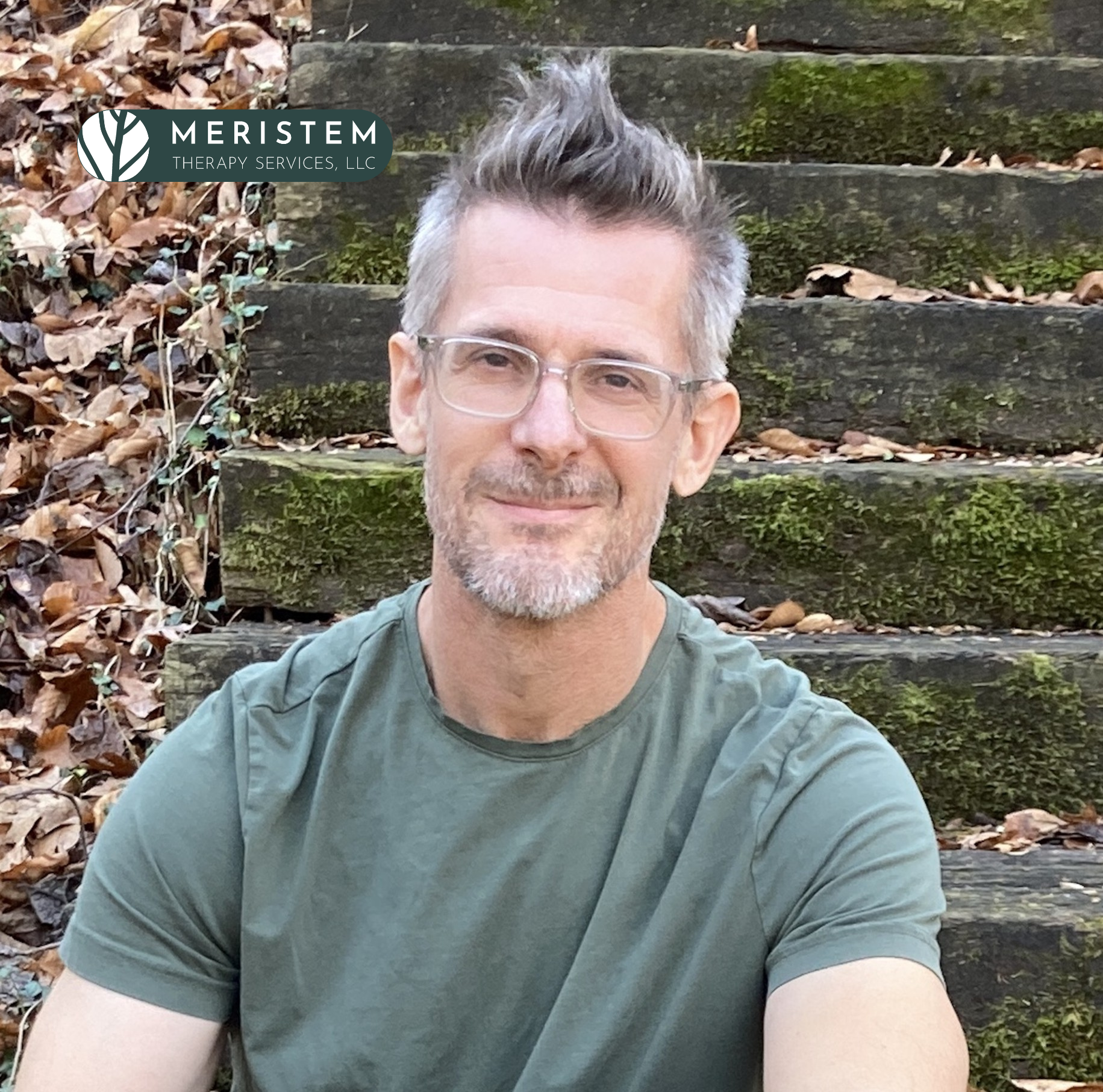 Picture of a light skinned man with short graying hair and glasses. He is wearing a t-shirt and sitting in front of steps that have moss on them and fallen leaves next to the steps.