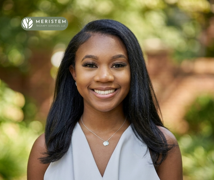 Headshot of young woman with dark skin, dark hair, and smiling. Background is blurry with green and browns, most likely outdoors.