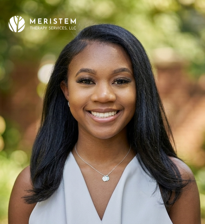 Young woman with dark skin and dark hair smiling with blurred green background outside.