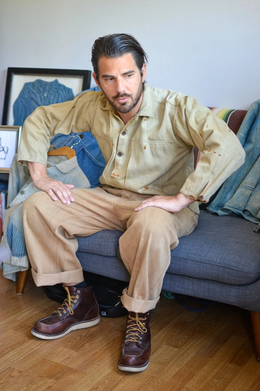 Un homme assis sur un canapé, portant une chemise beige sale, des pantalons beige, et des bottes en cuir marron, avec une expression pensive.
