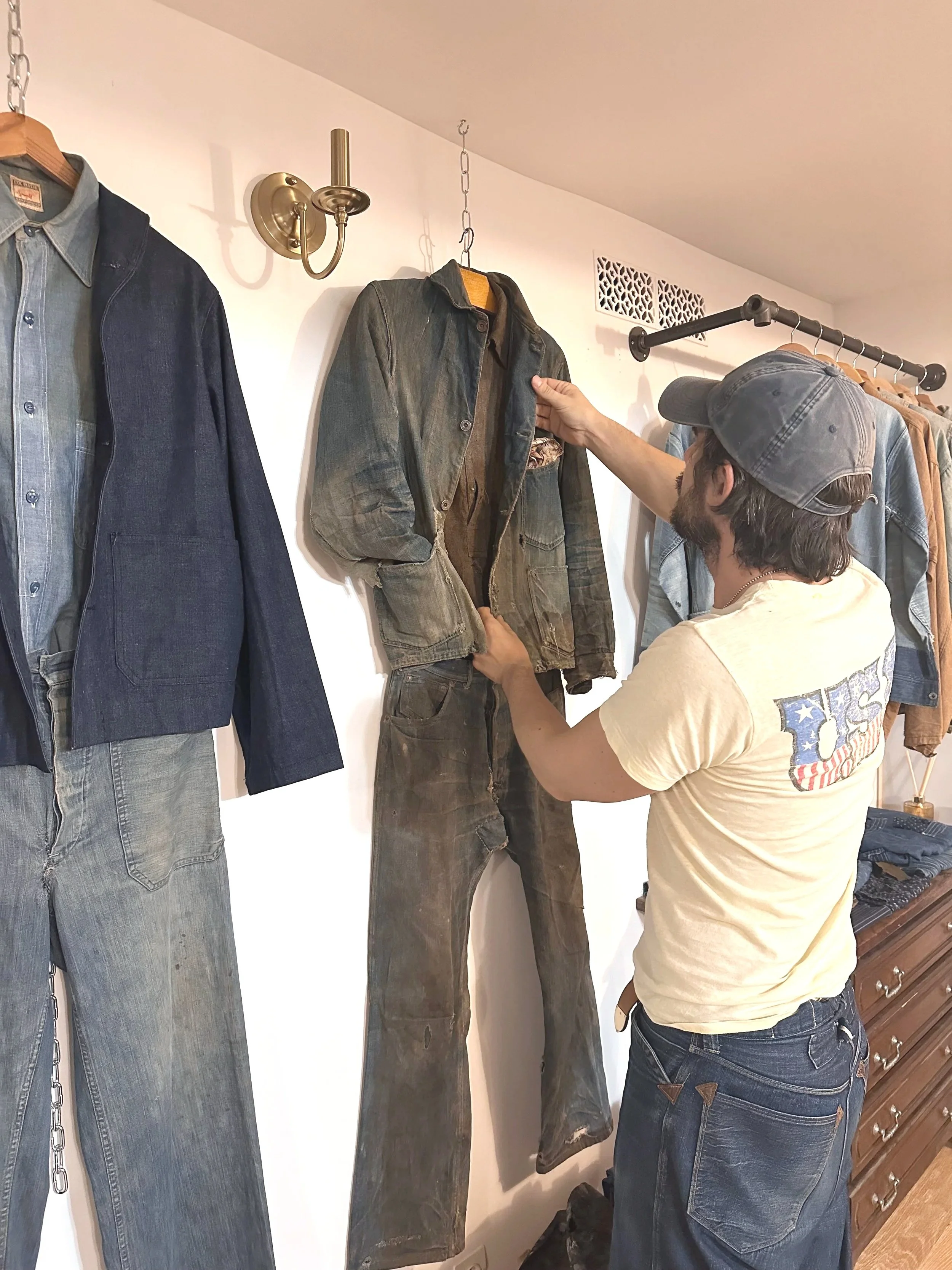 Un homme en casquette regarde des vêtements en denim et en cuir suspendus au mur dans une boutique. Il touche une veste en cuir.