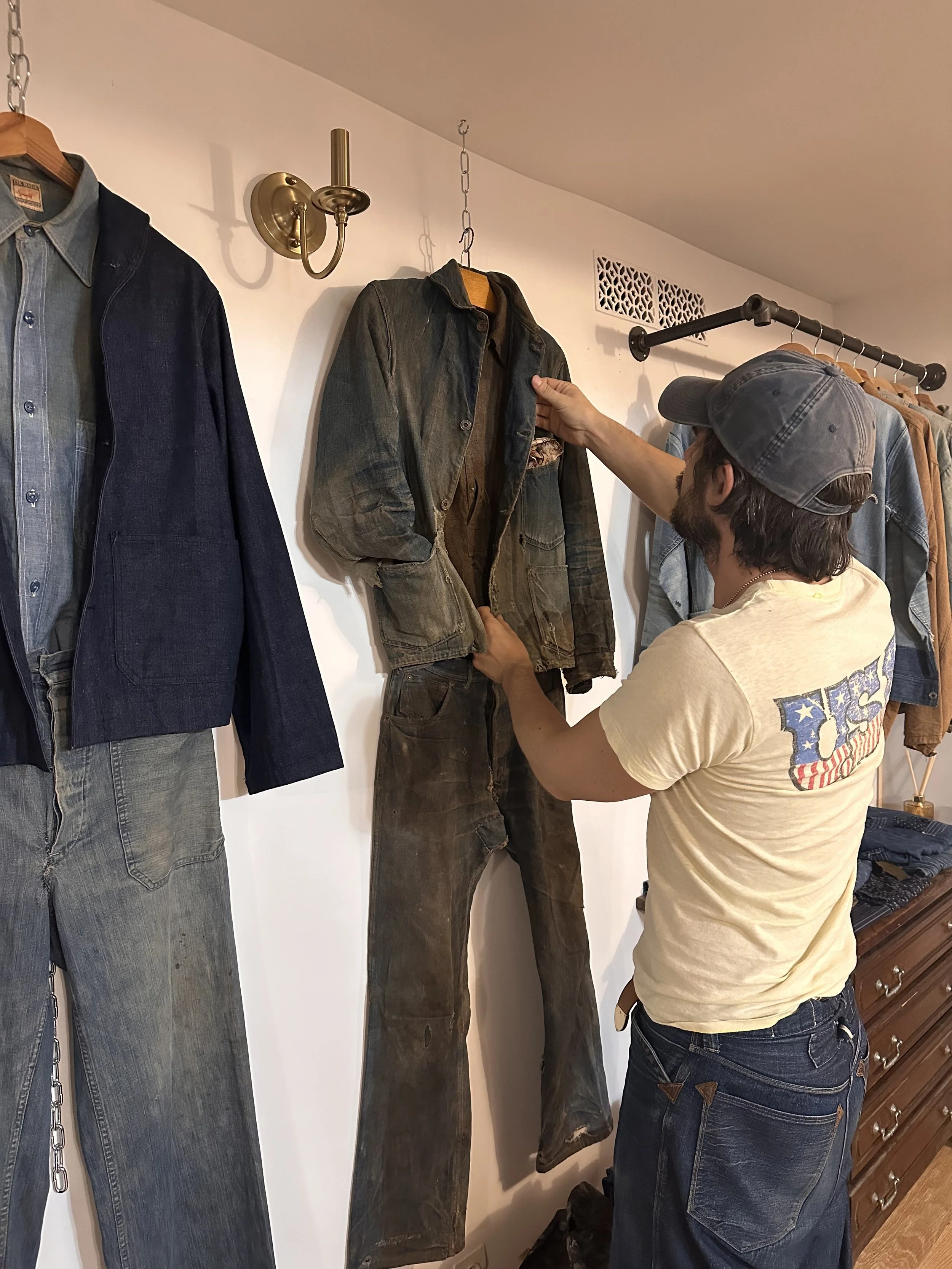 Un homme portant une casquette et un t-shirt clair essaye une veste en denim délavé dans un magasin de vêtements. La veste se trouve suspendue sur un mur blanc parmi d'autres vêtements, notamment des jeans et des chemises.