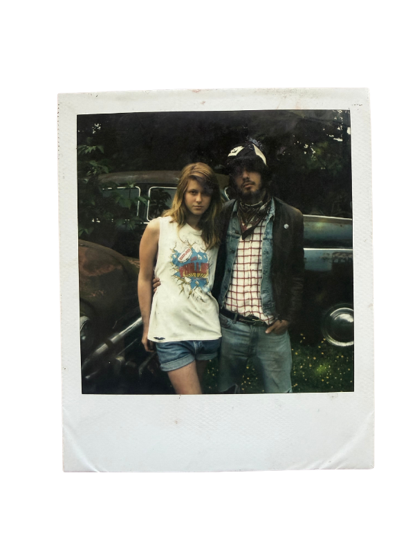 Une photo de style vintage montrant deux jeunes adultes, une femme et un homme, posant devant une voiture ancienne dans un environnement extérieur avec des arbres en arrière-plan.