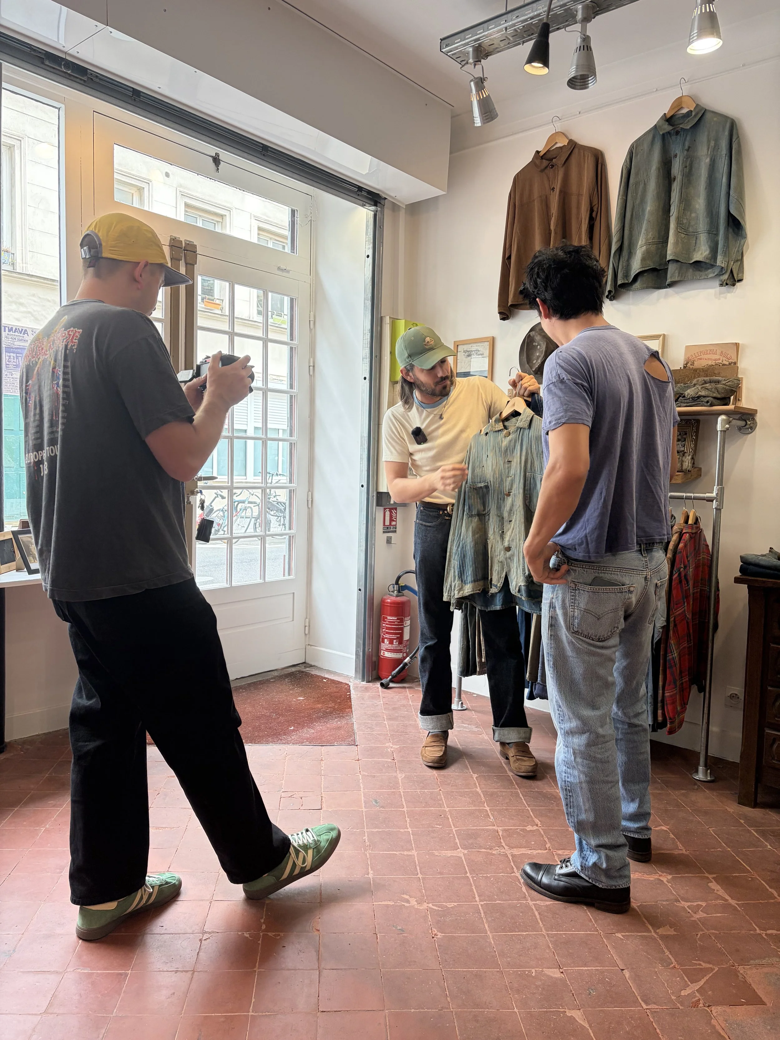 Quatre jeunes hommes dans un magasin de vêtements vintage, ils regardent un jean, l’un d’eux tient le vêtement et discute avec un autre, un troisième utilise son téléphone, des vêtements sont suspendus au mur et il y a une fenêtre avec la rue visible derrière eux.