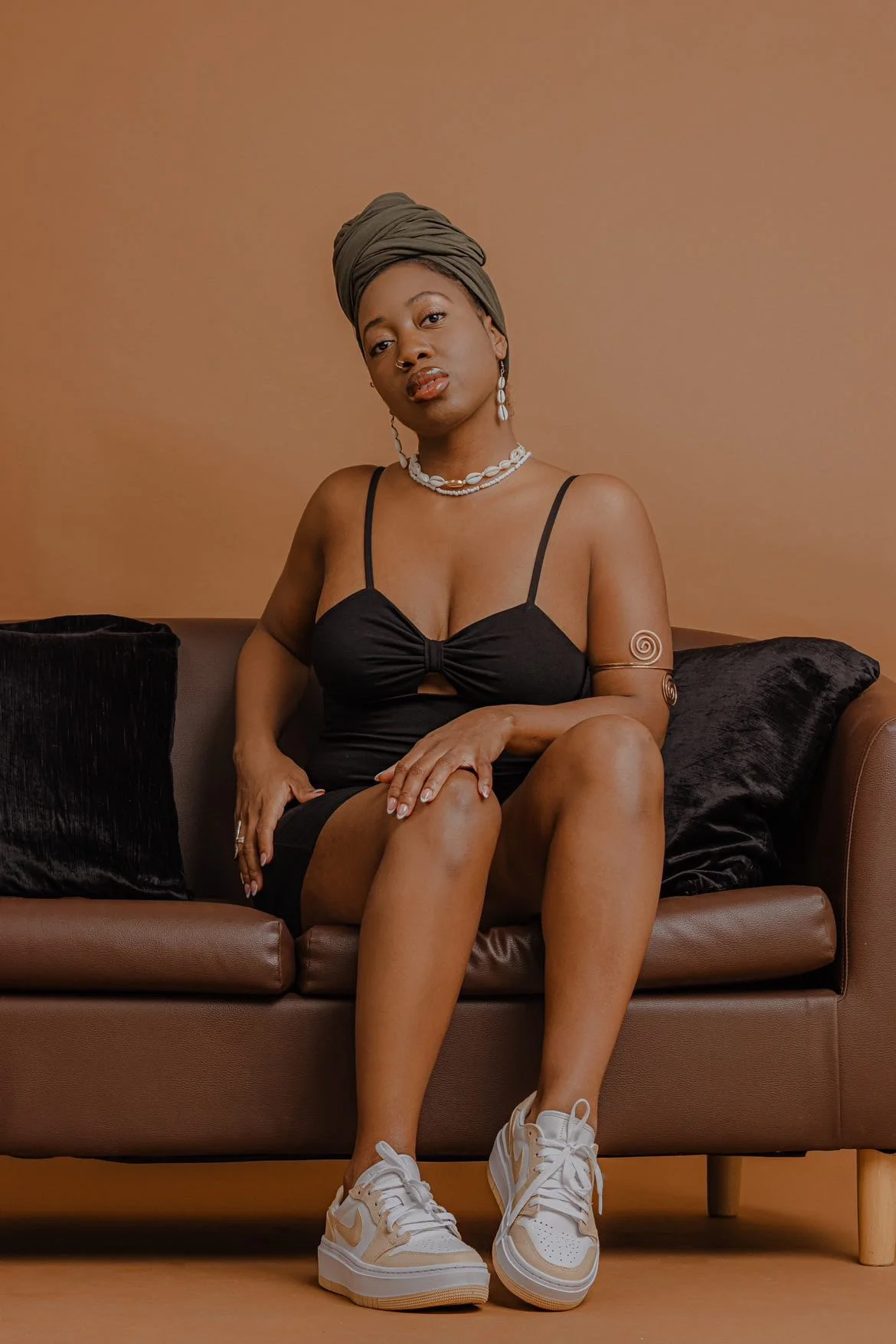 A woman sitting on a brown sofa against a warm brown background, wearing a black dress, white sneakers, and accessorized with jewelry and a headwrap.