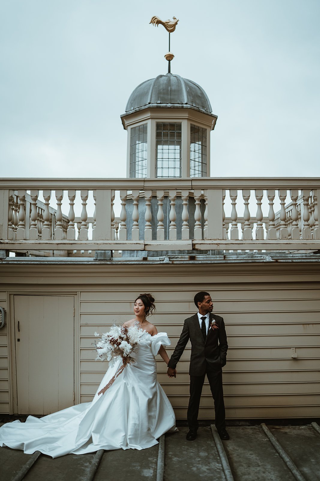 A bride and groom holding hands in front of a beige building with a cupola and weather vane on top. The bride is in a white wedding gown holding a bouquet, and the groom is in a black suit.