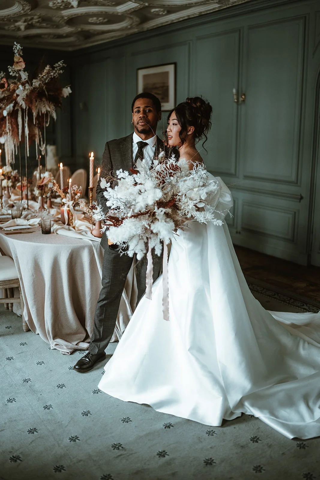 A bride in a white wedding gown standing next to a groom in a suit, holding a large bouquet of white flowers, in an elegant decorated room with a formal table setting and ornate ceiling.