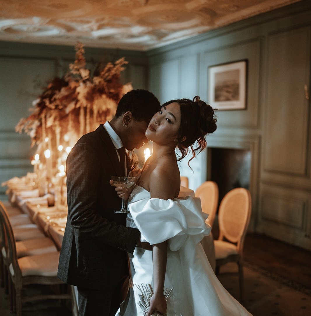 A couple at a wedding reception, the woman in a white off-shoulder dress holding a glass, the man in a dark suit, standing close together in an elegant room with a long table and a large floral centerpiece in the background.