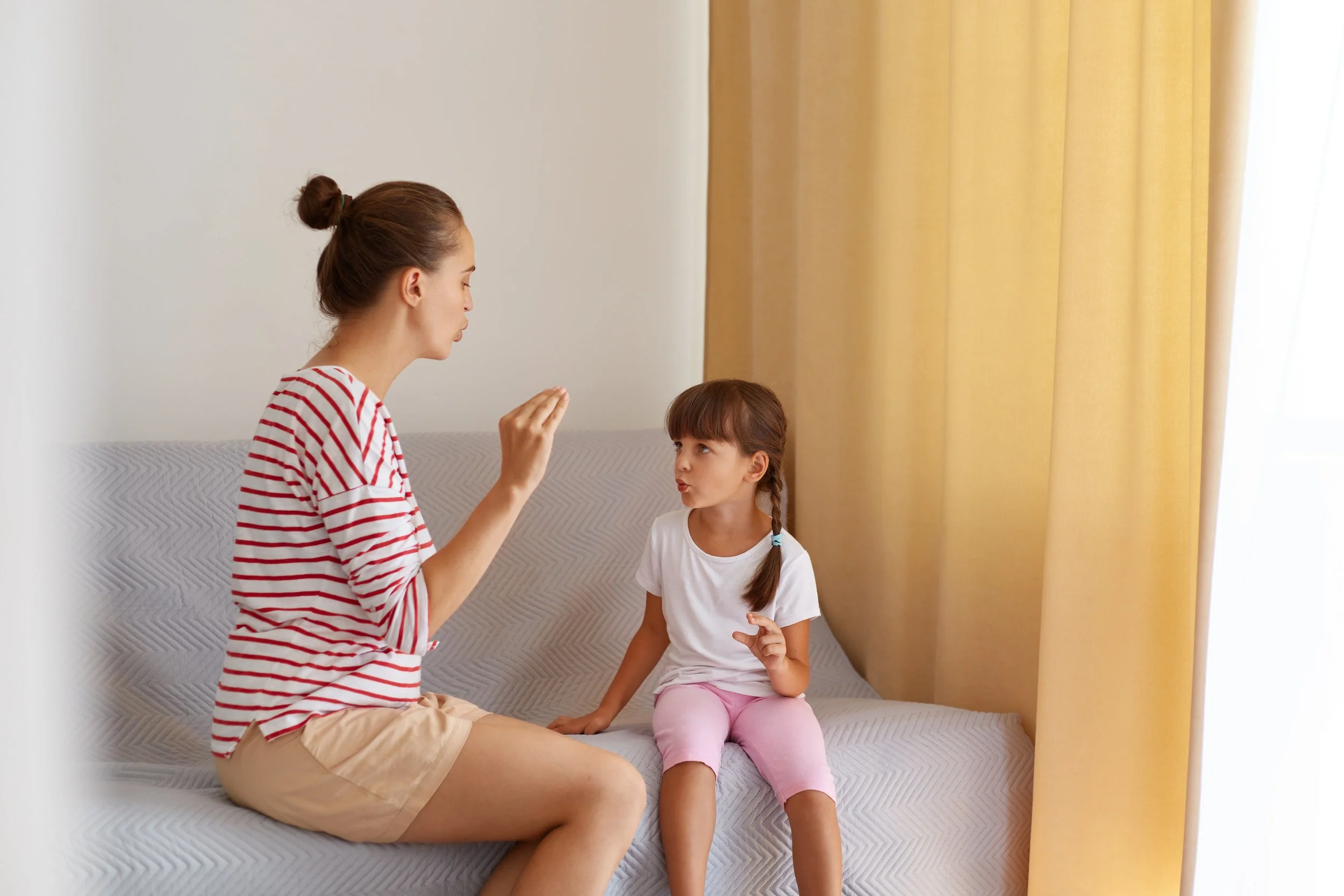 A woman and a young girl sit on a sofa, using sign language to communicate - Nurturing The Nest