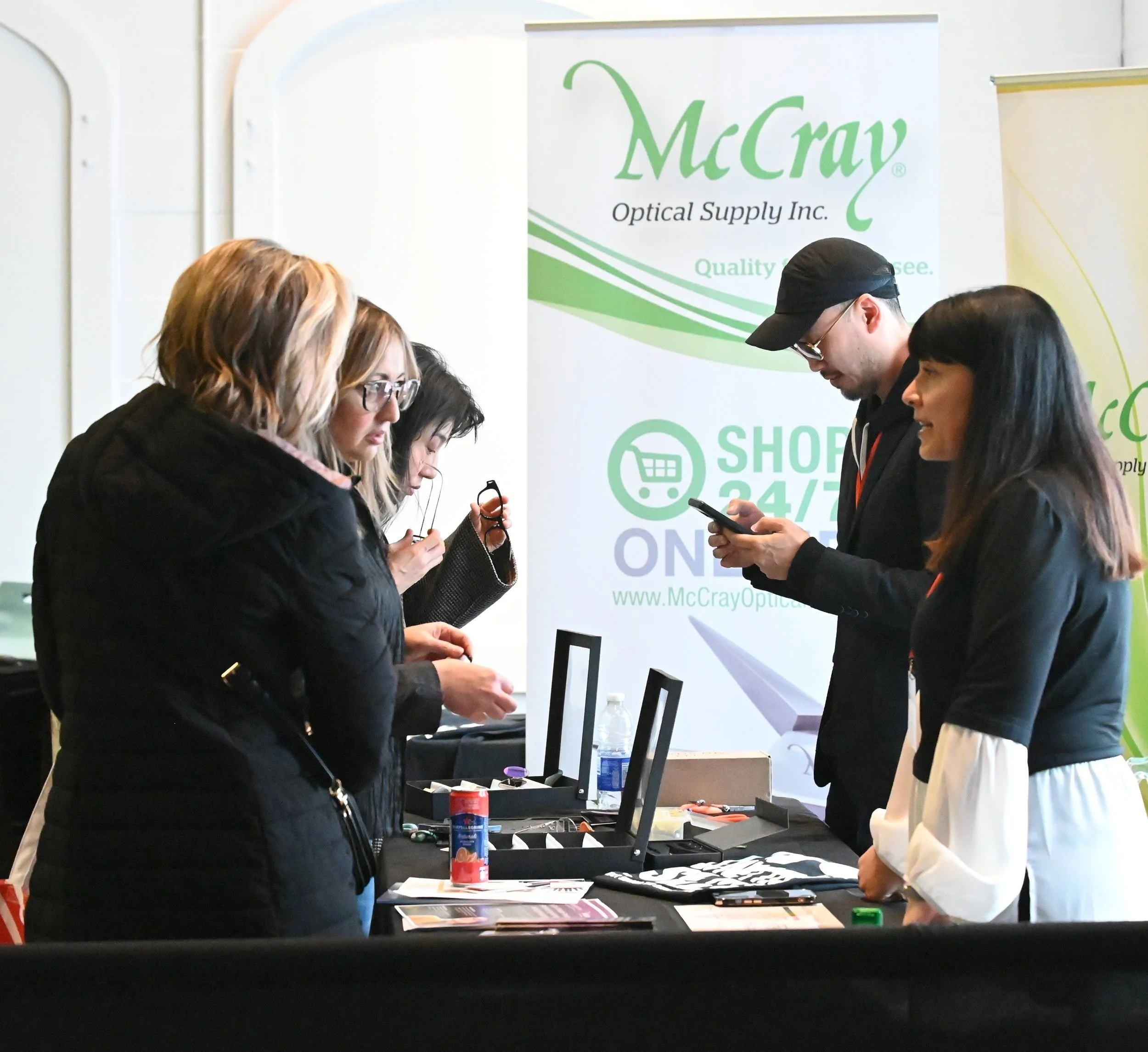 Group of five people shopping for glasses at an event booth with a McCray Optiical Supply Inc. banner in the background.