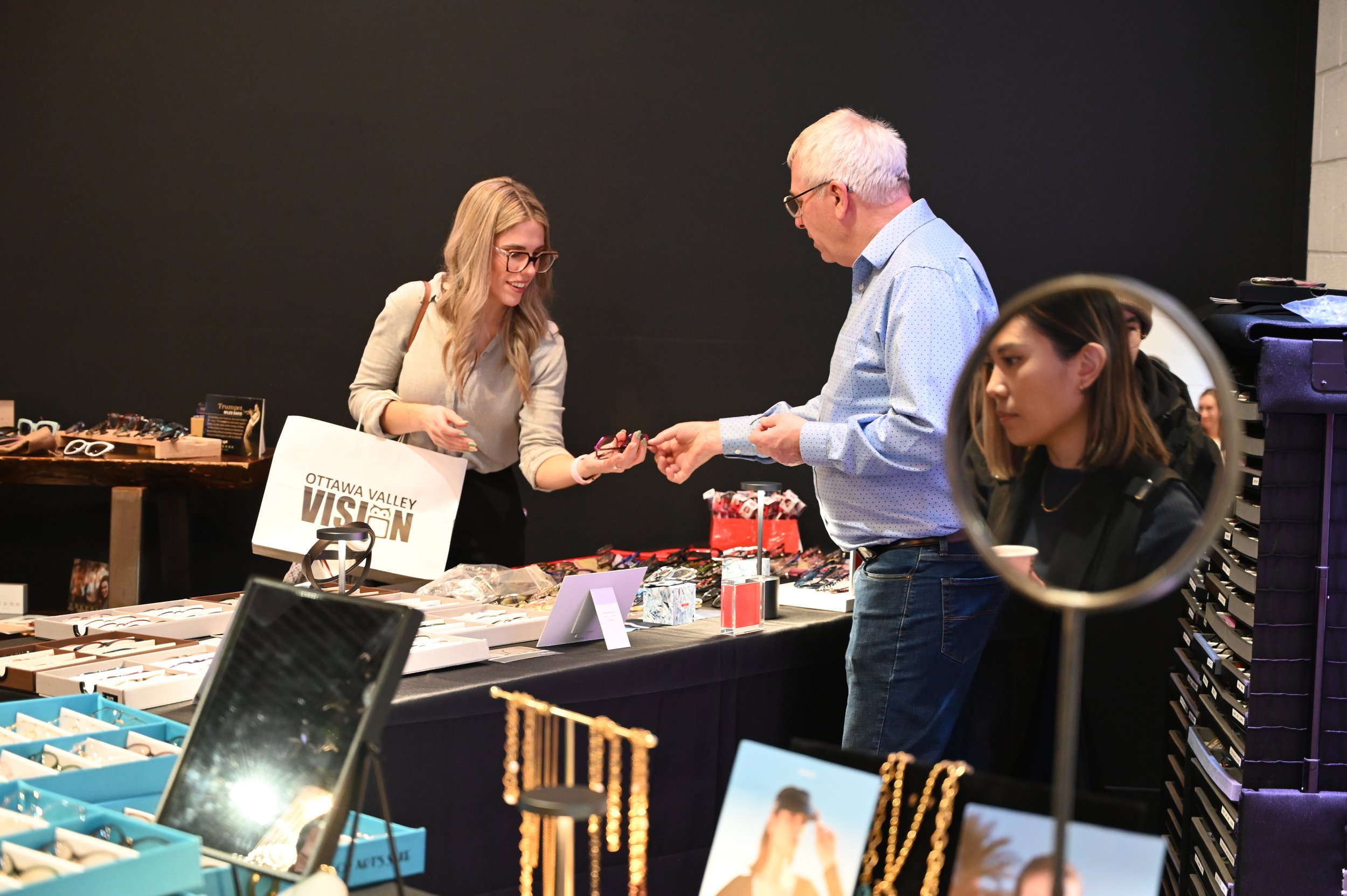 A woman shopping at a jewelry booth, showing her purchase to an older man, with another woman sitting nearby.
