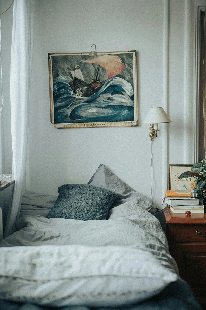 A bedroom with a bed and a nightstand, a painting of a boat in rough seas hangs above the bed, a wall-mounted lamp is on the right side of the image, and a window with light curtains is on the left side.
