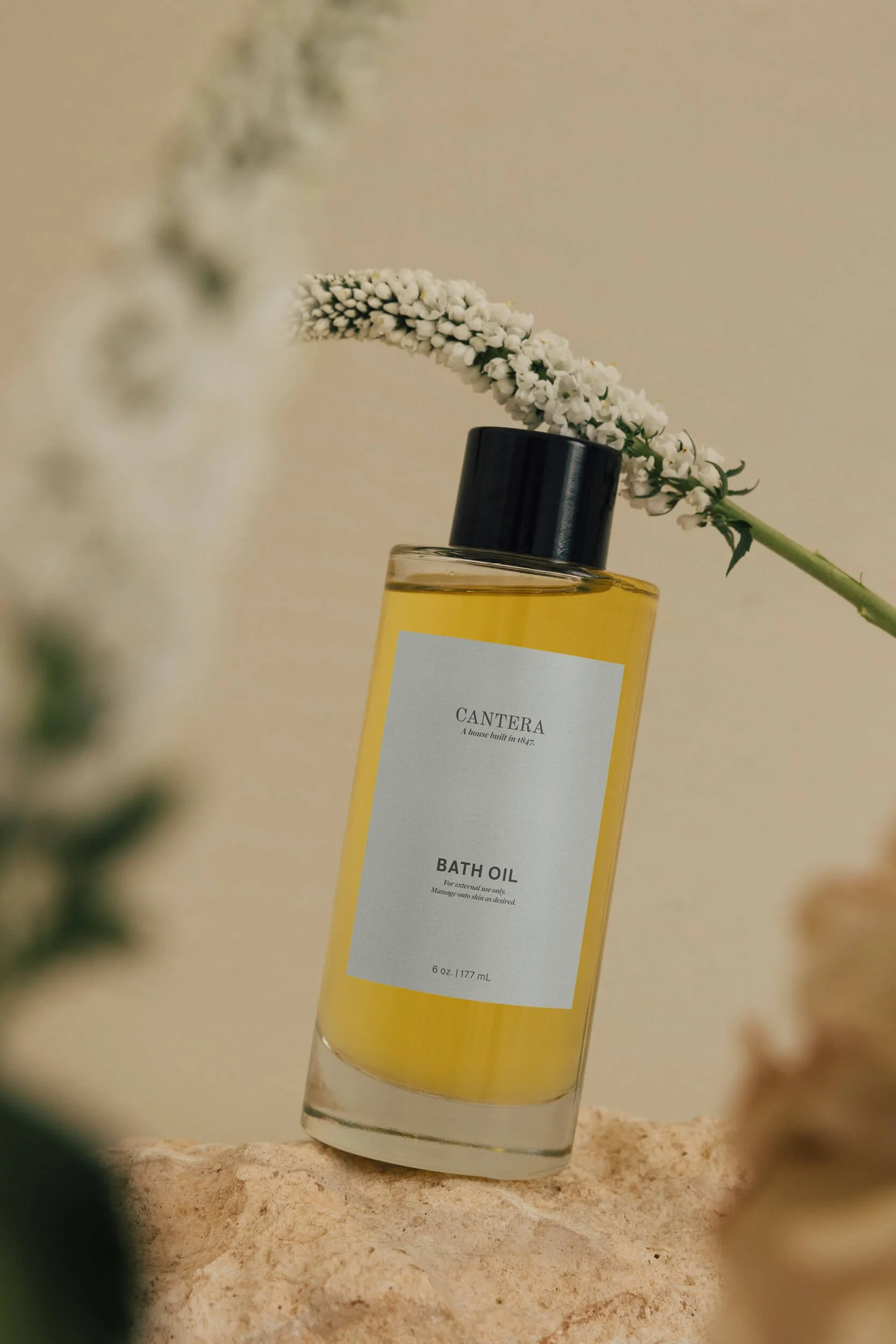 A bottle of bath oil with a white label and black cap, placed on a sandy surface with a white flower resting on top of the bottle.