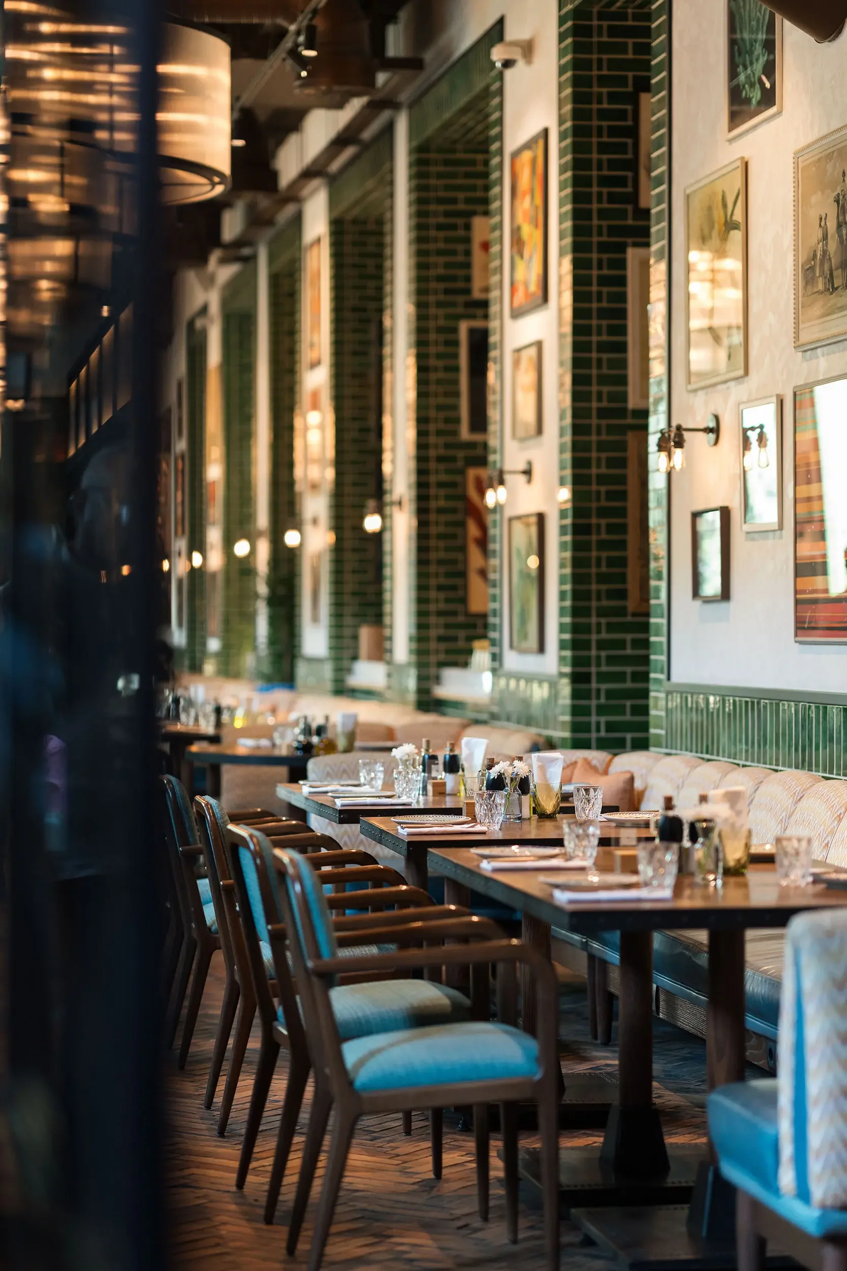 Interior of a restaurant with arranged tables, wooden chairs, and framed artwork on the walls. Green tiled accents and wall-mounted lamps are visible.