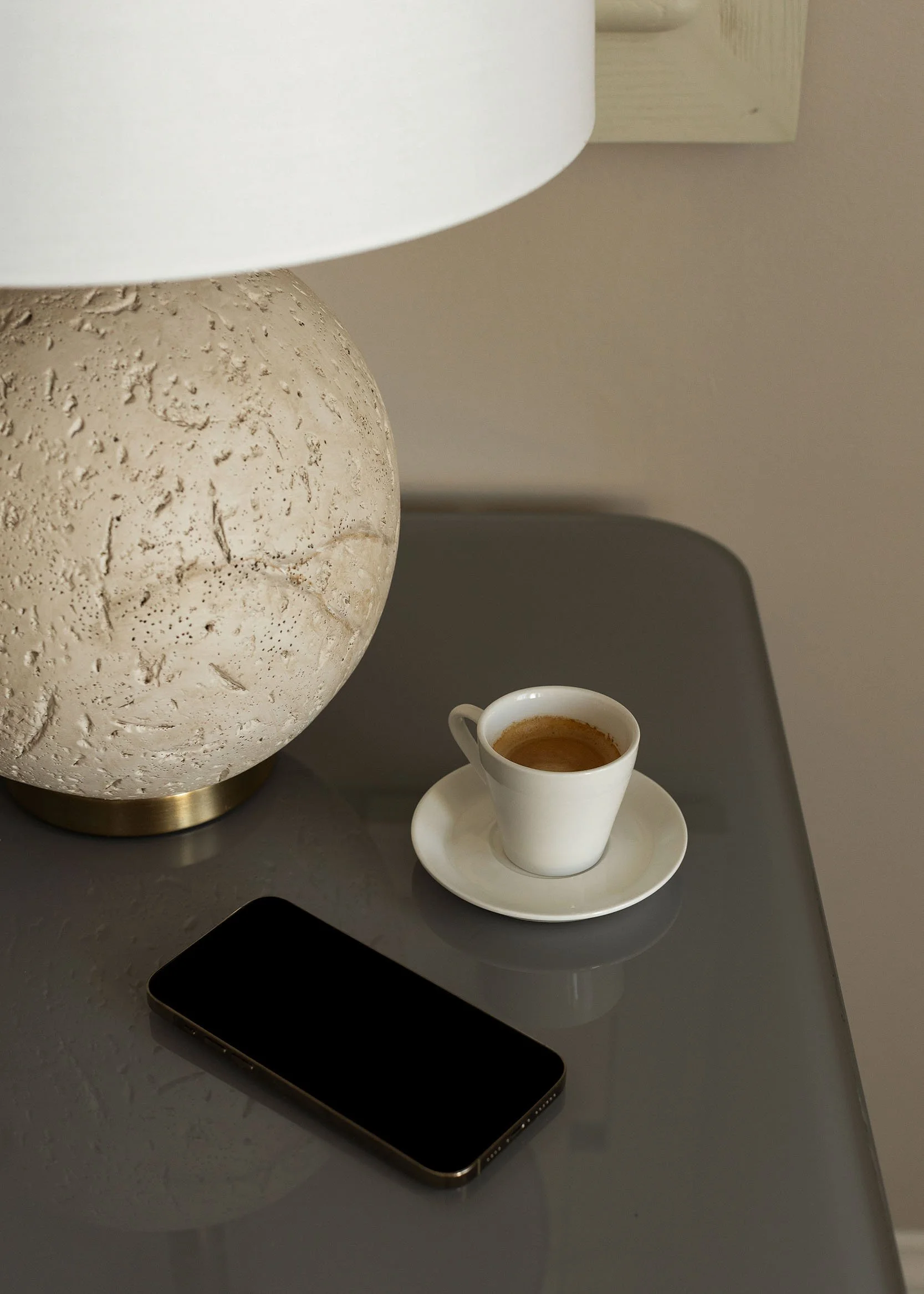 A beige textured lamp, a white paper cup filled with espresso on a white saucer, a black smartphone, and part of a grey table surface.