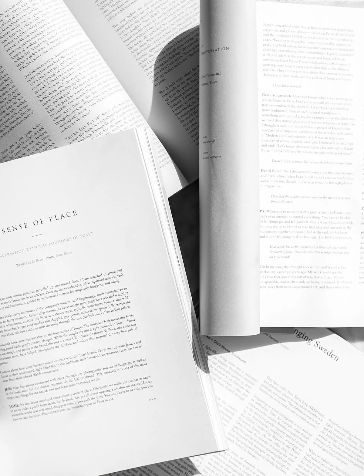 A collection of open books and magazines in black and white, with some pages overlapping each other, displaying text and titles like "A Sense of Place" and "Conversation with the Founders of Toast."