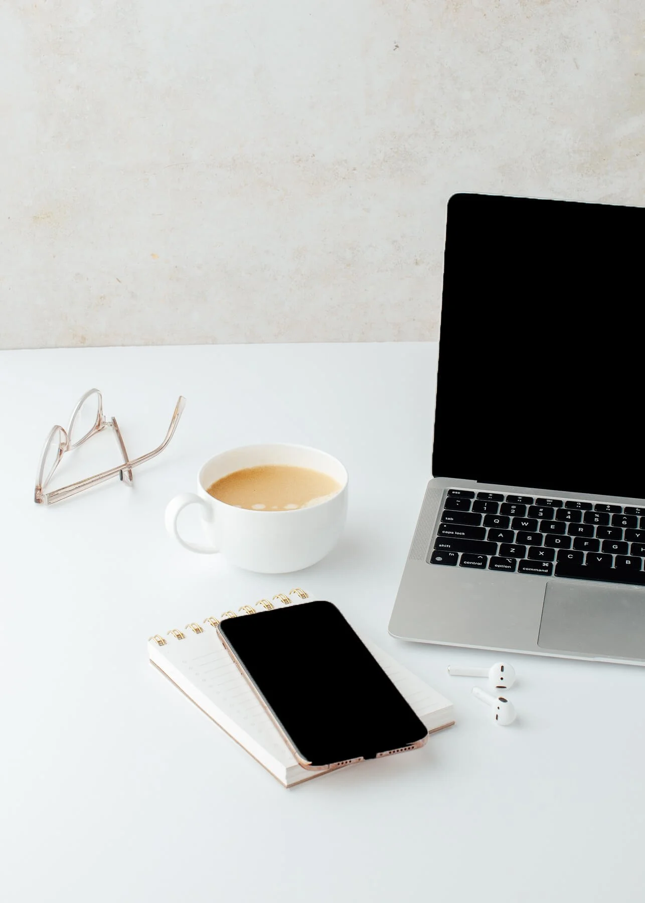 Desk workspace with a laptop, smartphone, notebook, wireless earbuds, a cup of coffee, glasses, and a notepad on a white surface with a neutral wall background.