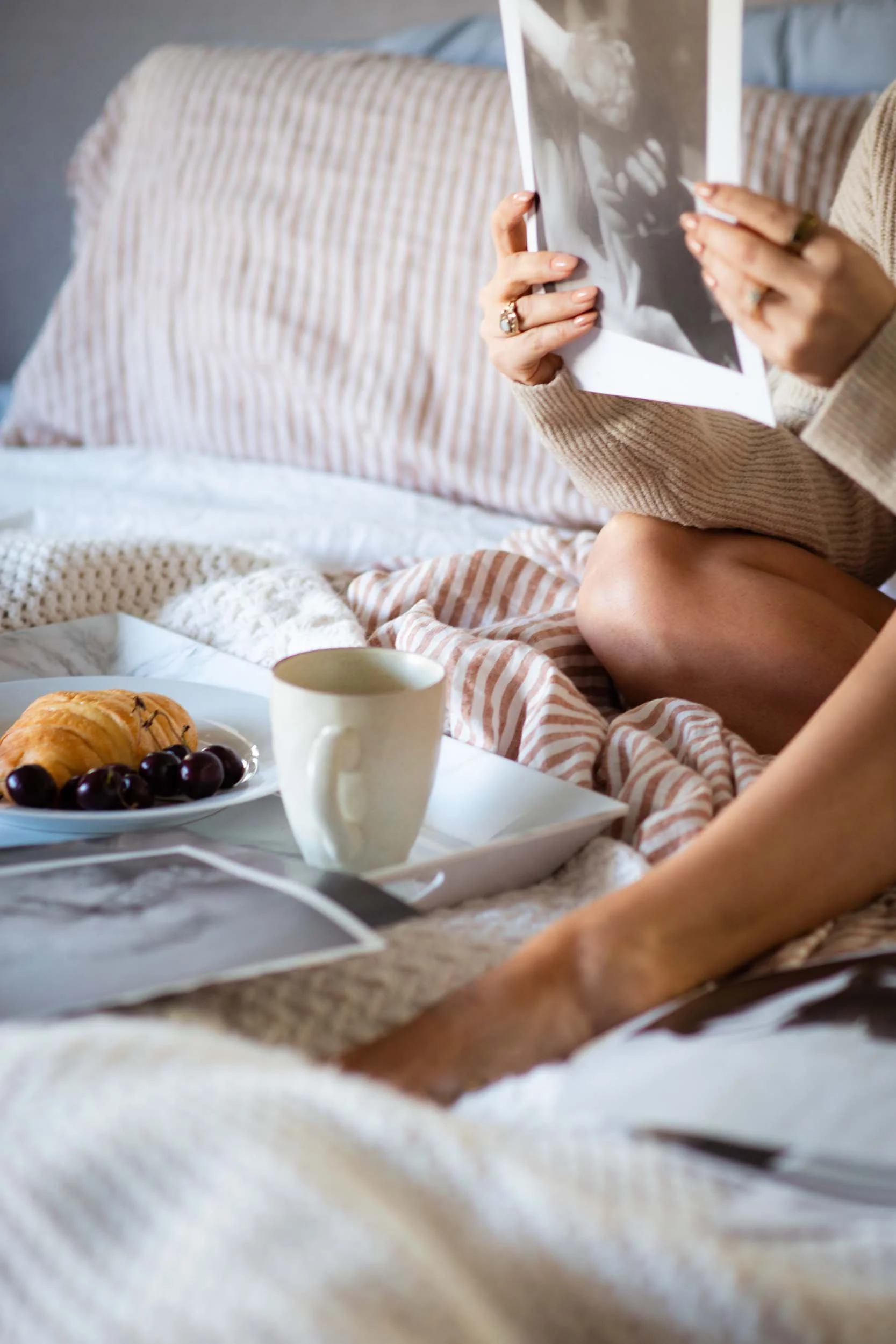 A person holding a black-and-white photograph, sitting on a bed with striped bedding. A plate of croissants and grapes, along with a white mug, are on a tray in the foreground.