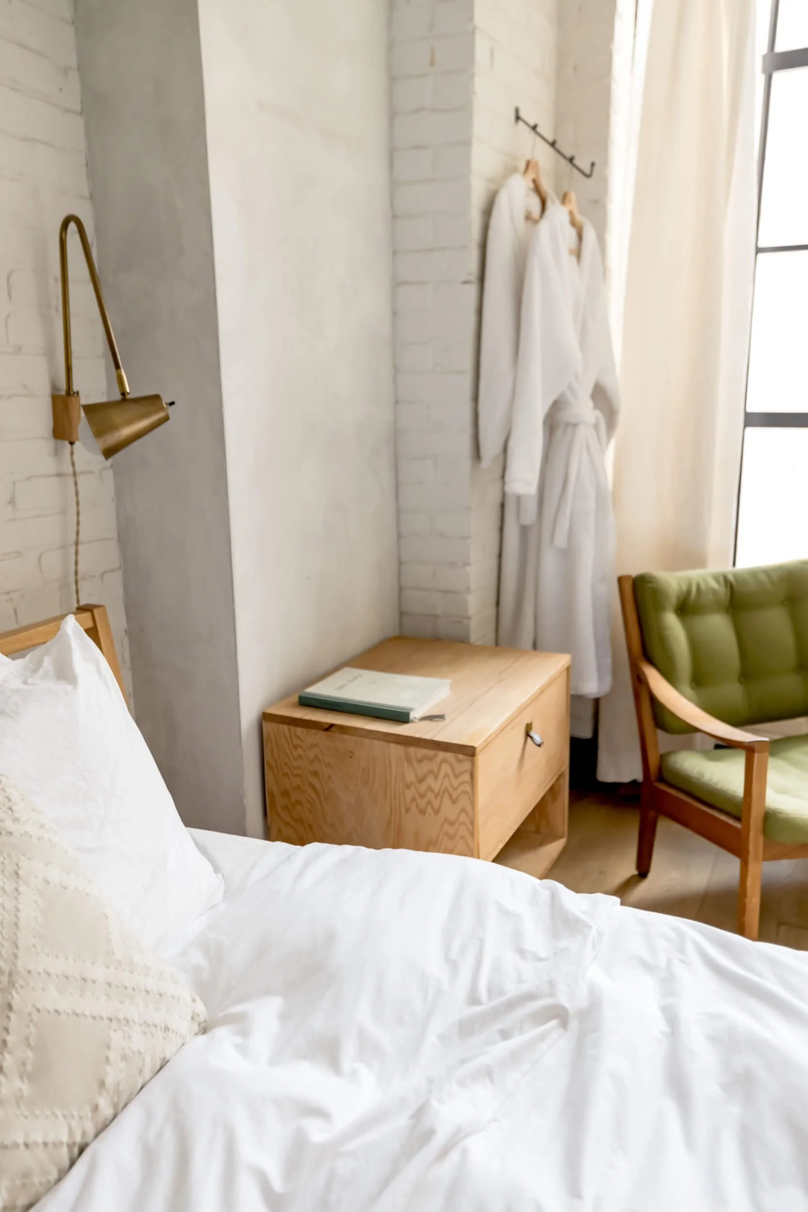 A cozy bedroom corner with a bed, a wooden side table, a green armchair, a wall-mounted gold reading lamp, white towels hanging on hooks by a window, and white curtains.