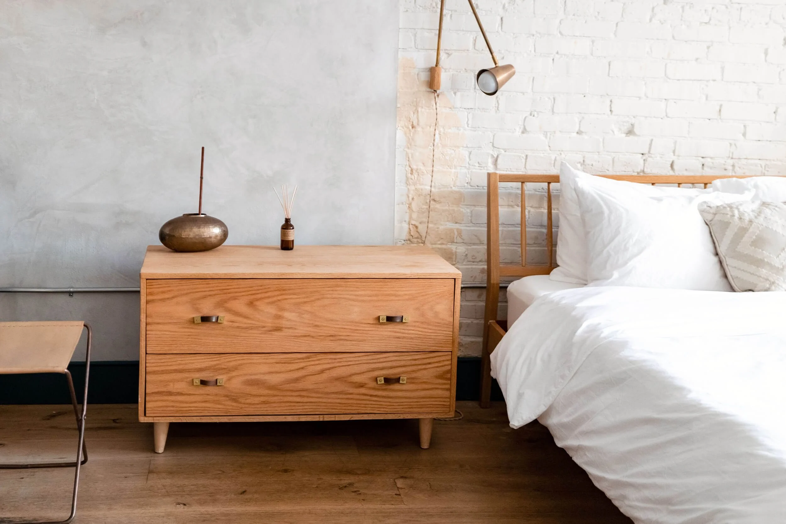 A bedroom with a wooden bed frame with white bedding, a wooden dresser with two drawers, a metallic-colored décor item and a reed diffuser on top, a wall-mounted reading lamp, a white brick wall, and a concrete wall.