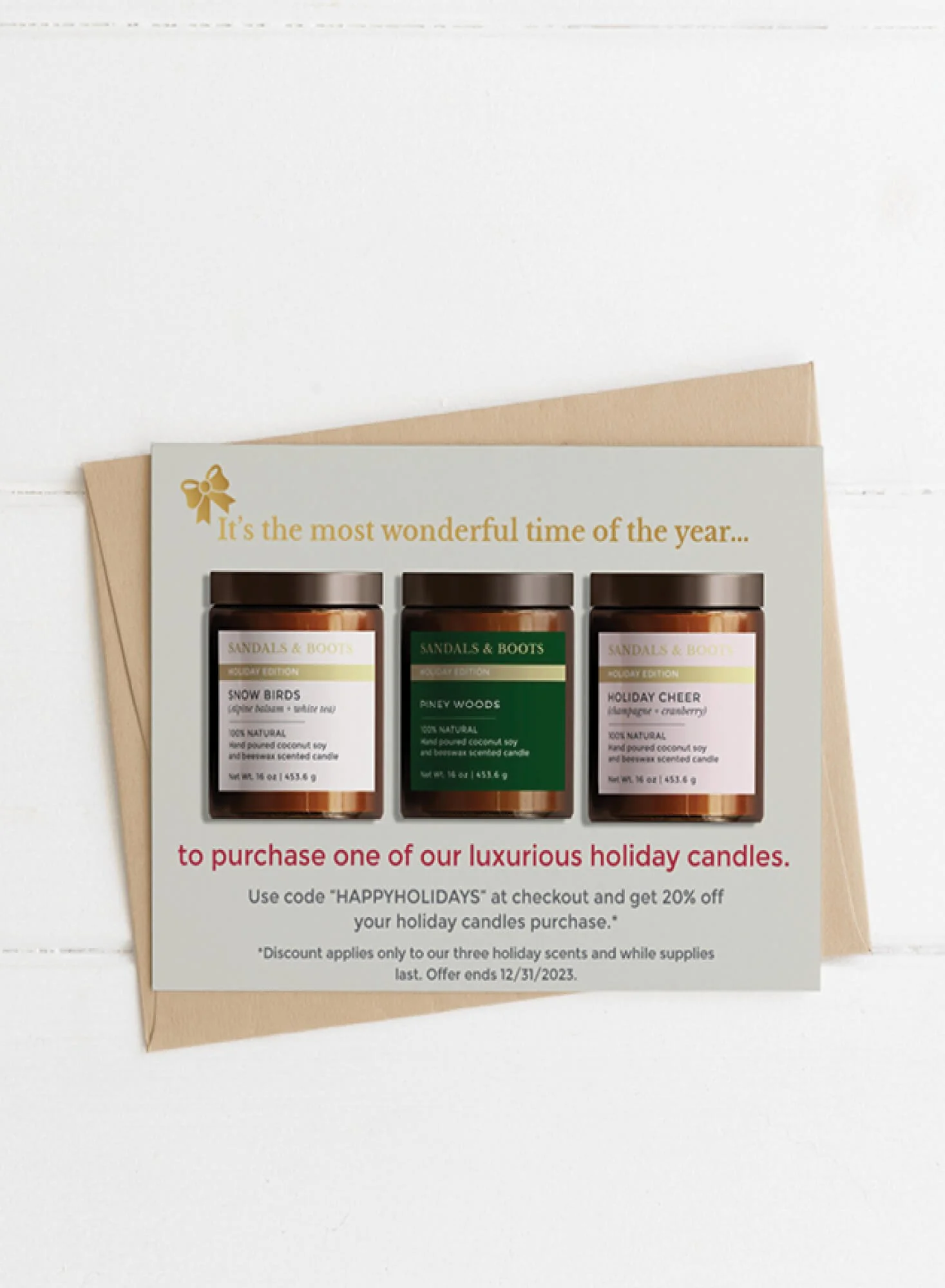 Sandals & Boots holiday promotional postcard displayed against a kraft envelope on a white shiplap background. The sage green card features a gold bow illustration, three Holiday Edition candles (Snow Birds, Piney Woods, and Holiday Cheer), and a dis