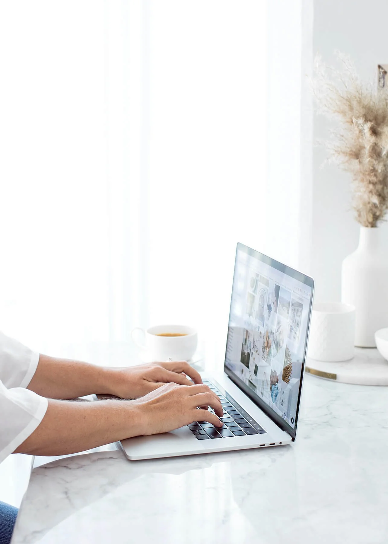 Person working on a MacBook laptop at a white marble desk, with a coffee cup and a vase with dried flowers nearby, in a bright, minimalistic room.