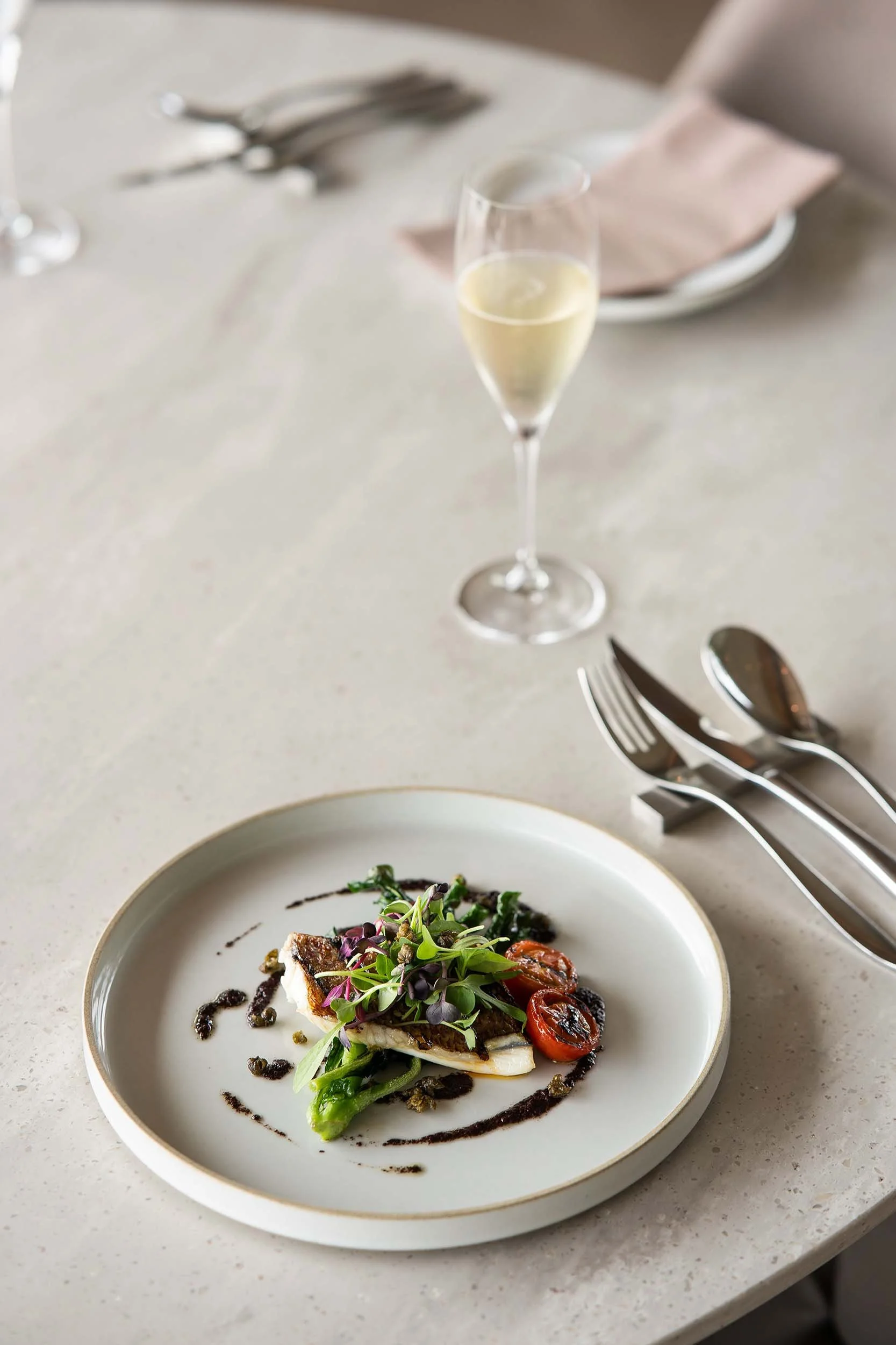 A plated gourmet dish featuring a piece of cooked fish, topped with microgreens and cherry tomatoes, garnished with a drizzle of dark sauce, on a white plate with a gold rim. In the background, a champagne flute filled with champagne and a dining setting with silverware, a wine glass, and a napkin on a light-colored table are visible.