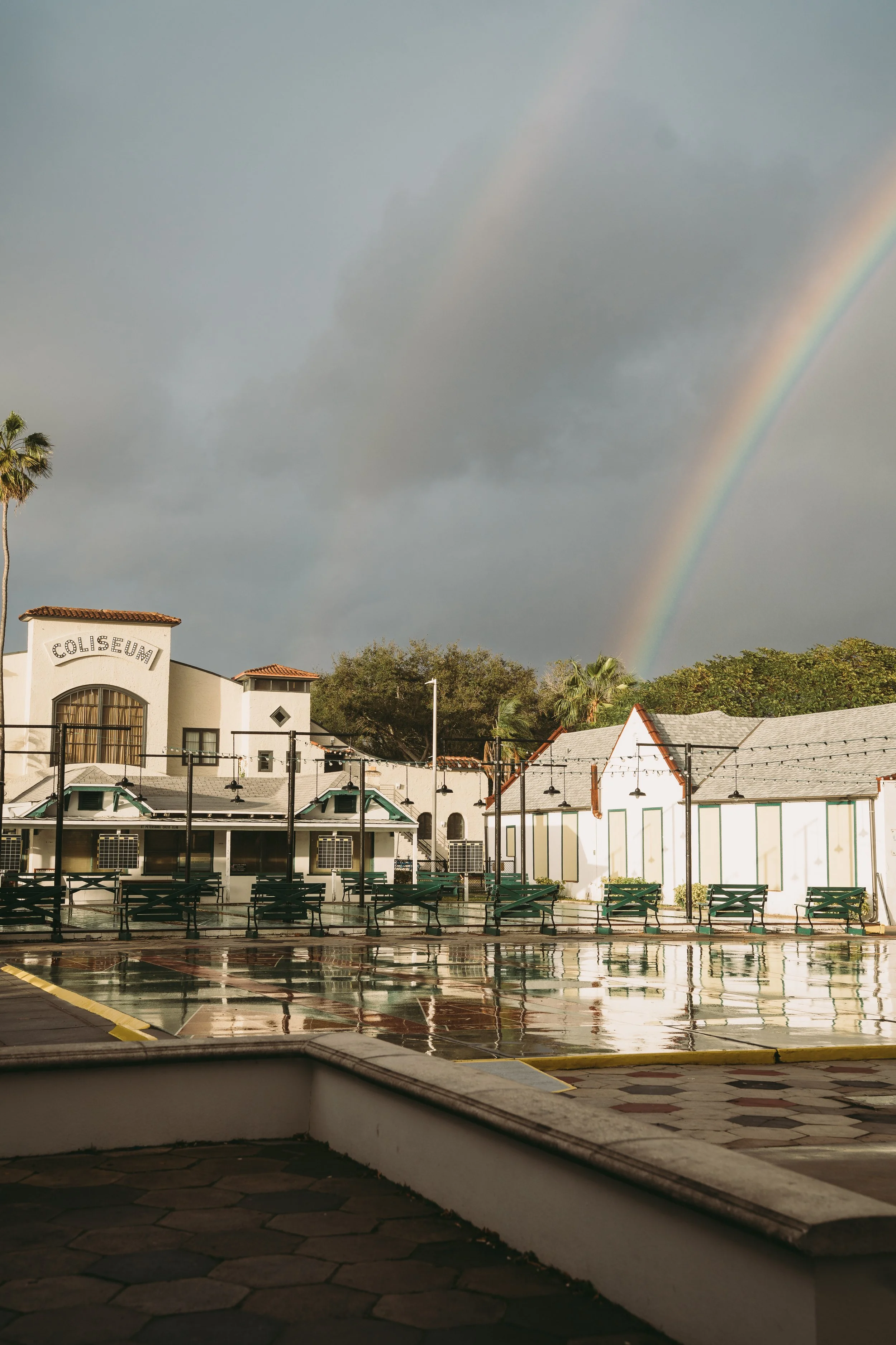 St. Pete Florida wedding photographer capturing shuffleboard club photo, 'Coliseum' on one building, wet pavement reflecting the surroundings, a rainbow in the cloudy sky, palm trees, and empty benches - Jennifer Elizabeth Studios
