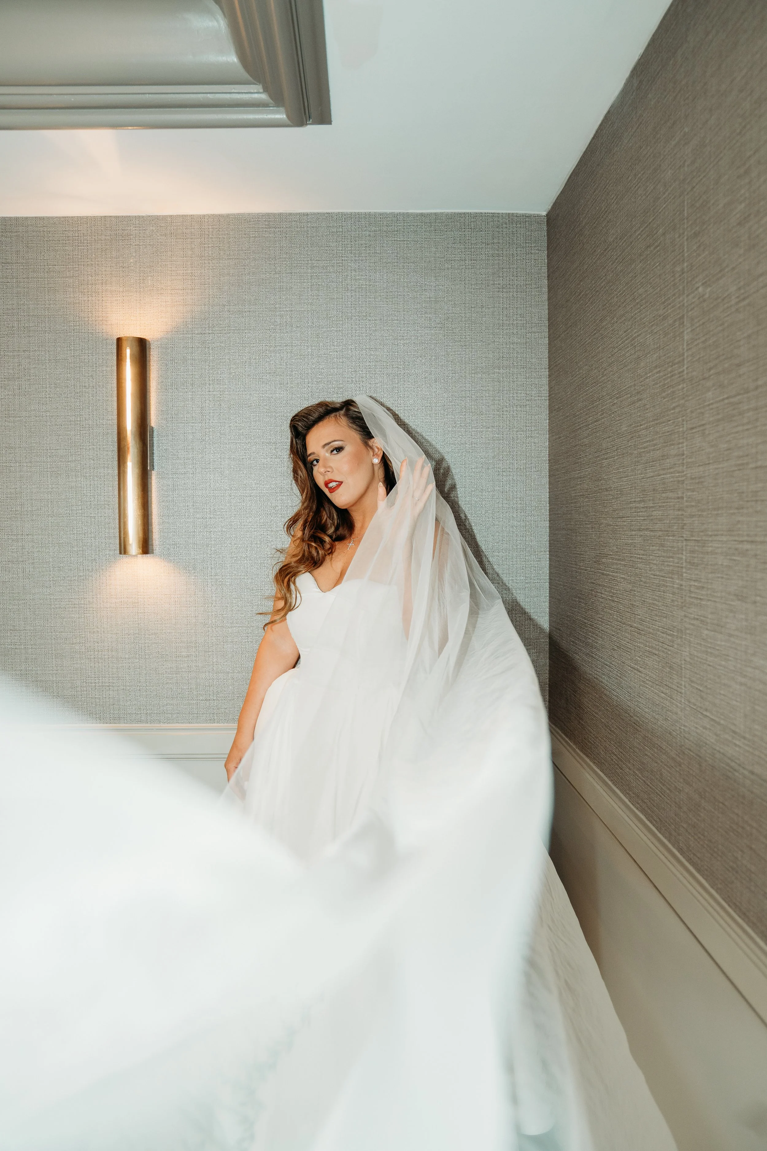 St. Petersburg Florida wedding photographer capturing Vinoy Hotel – Jennifer Elizabeth Studios
A bride in a white wedding dress with a veil posing in a room with textured gray walls and a sconce light.