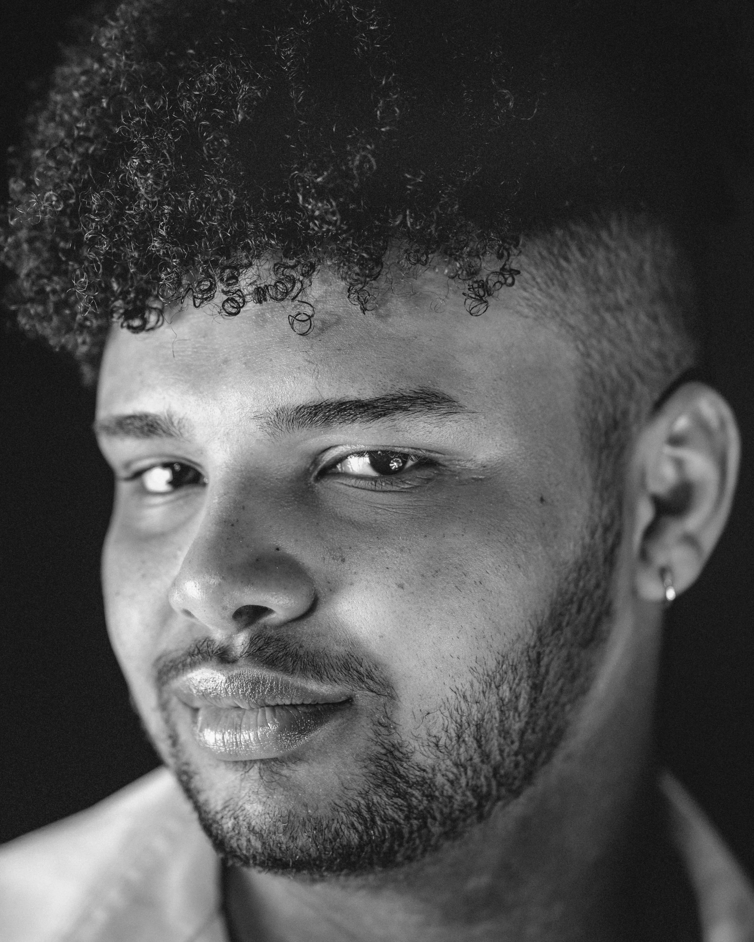 Close-up black and white portrait of a man with short curly hair on top and shaved sides, facial hair, and an earring