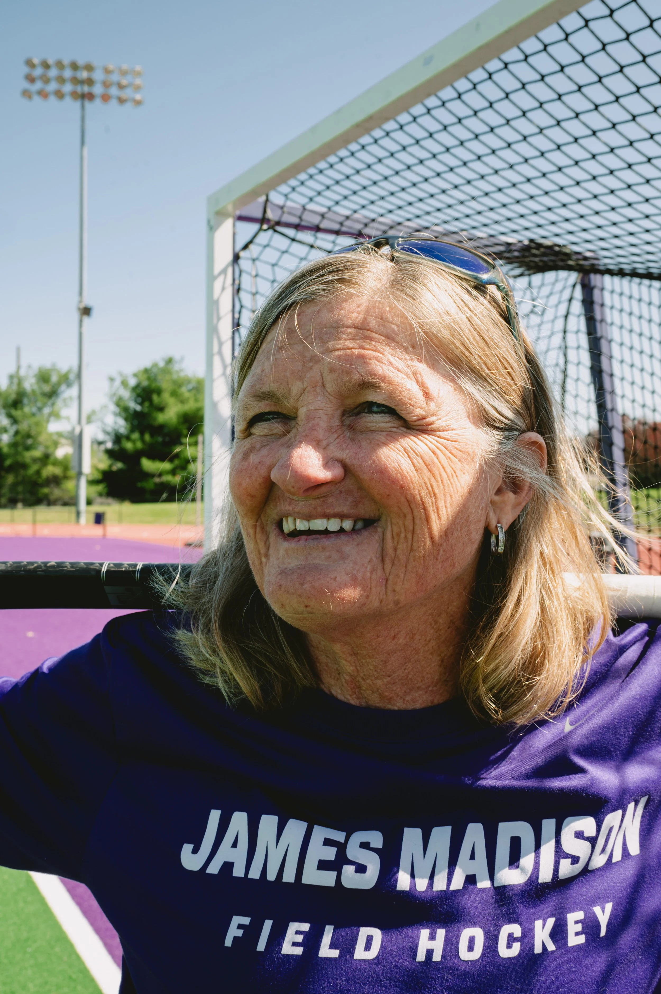 A smiling woman with sunglasses on her head, wearing a purple James Madison Field Hockey shirt, standing near a field hockey goal on an outdoor field.