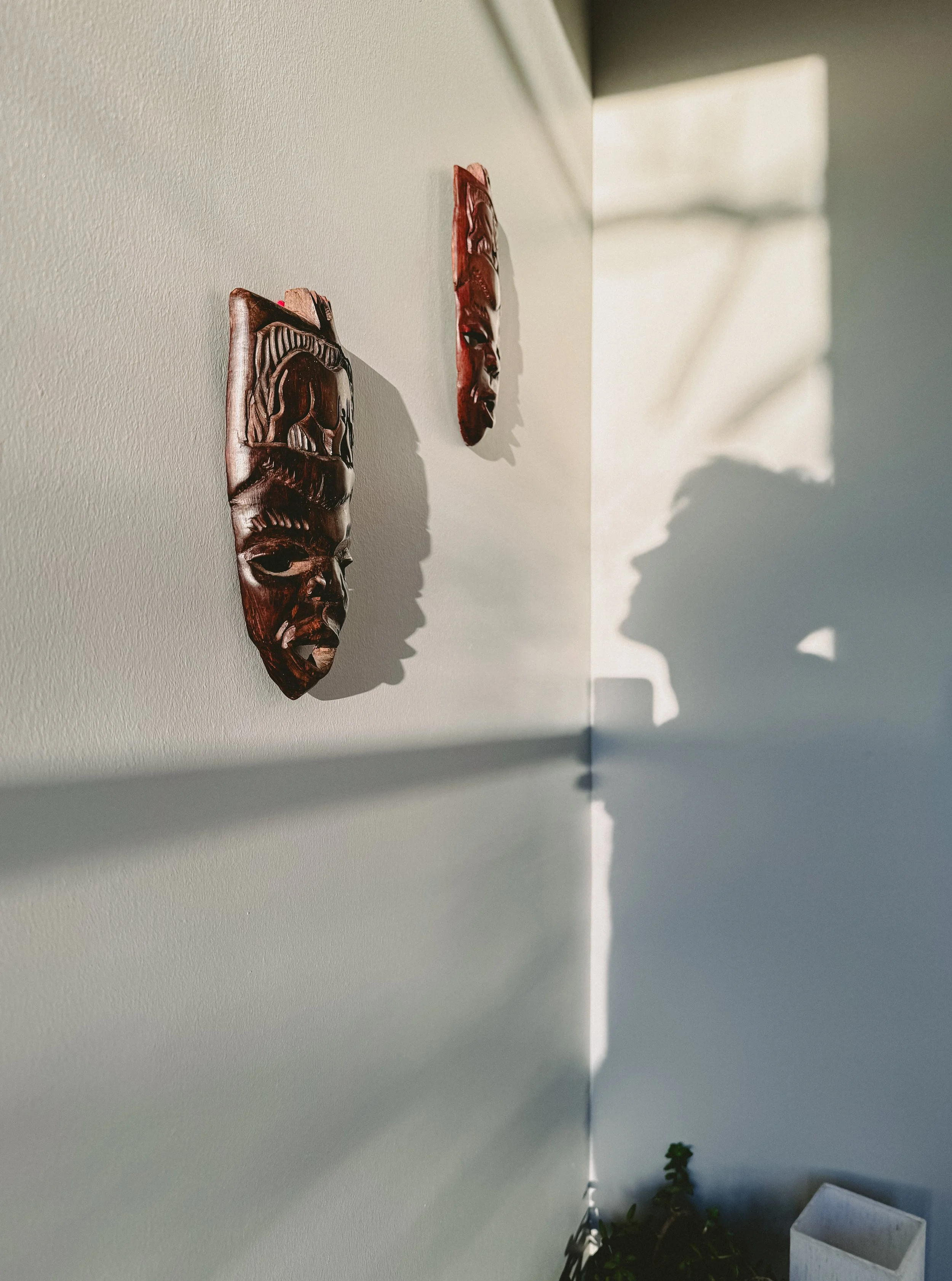 Two decorative wooden masks hanging on a light-colored wall with shadows cast by sunlight, and a small plant pot at the bottom.