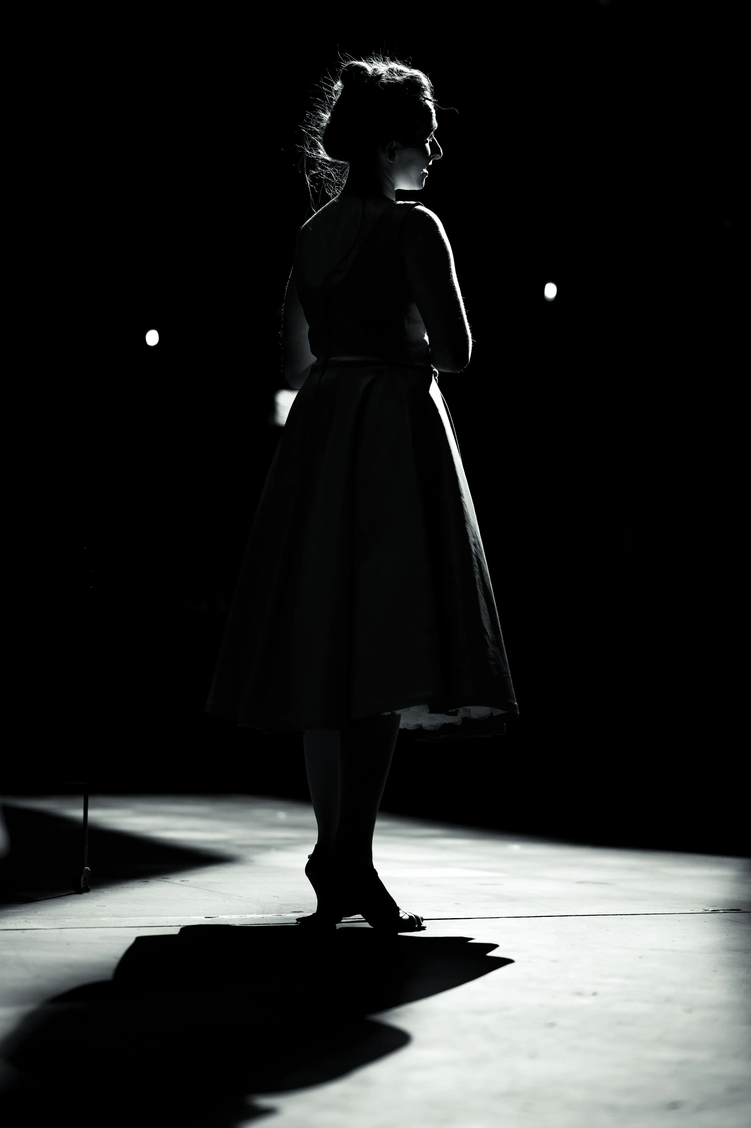 Black and white silhouette of a woman standing on stage with dramatic lighting, wearing a vintage dress.