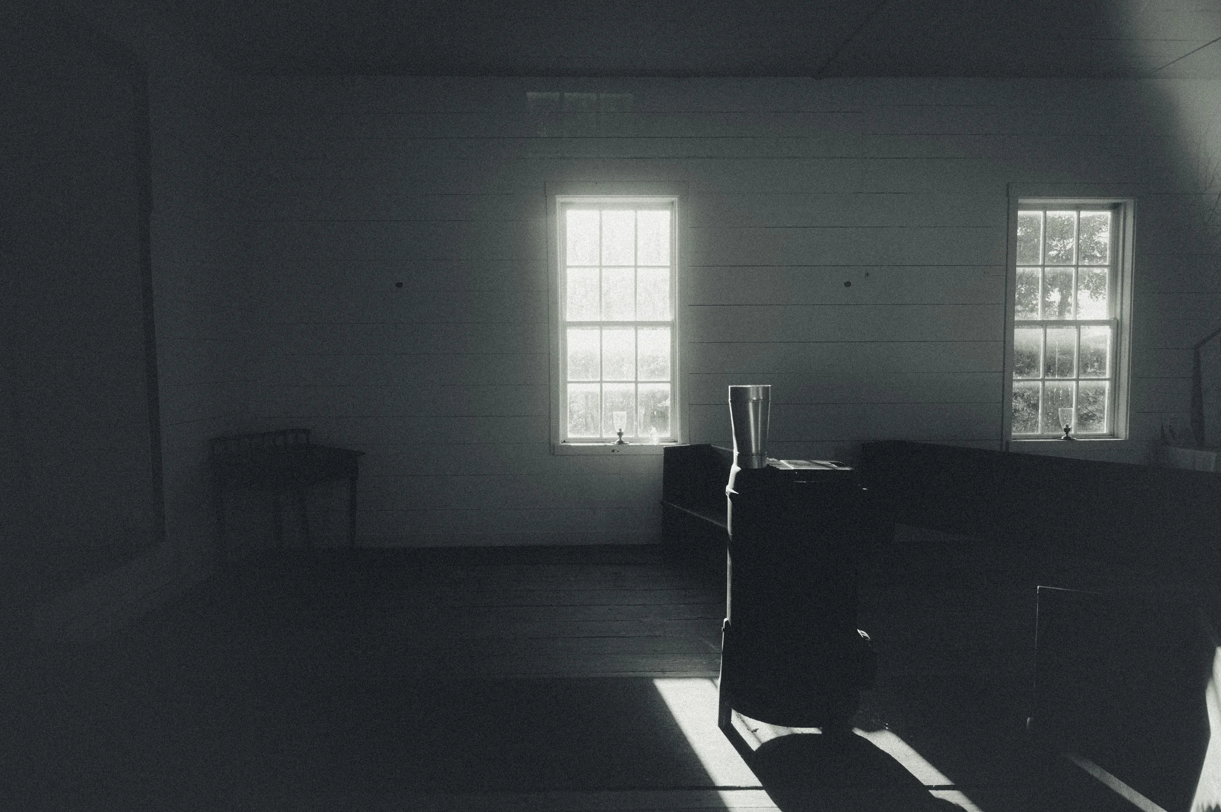 A dimly lit room with two windows allowing natural light, a desk in the center with a tall glass of water and some papers, and a chair in front of the desk, all in black and white.