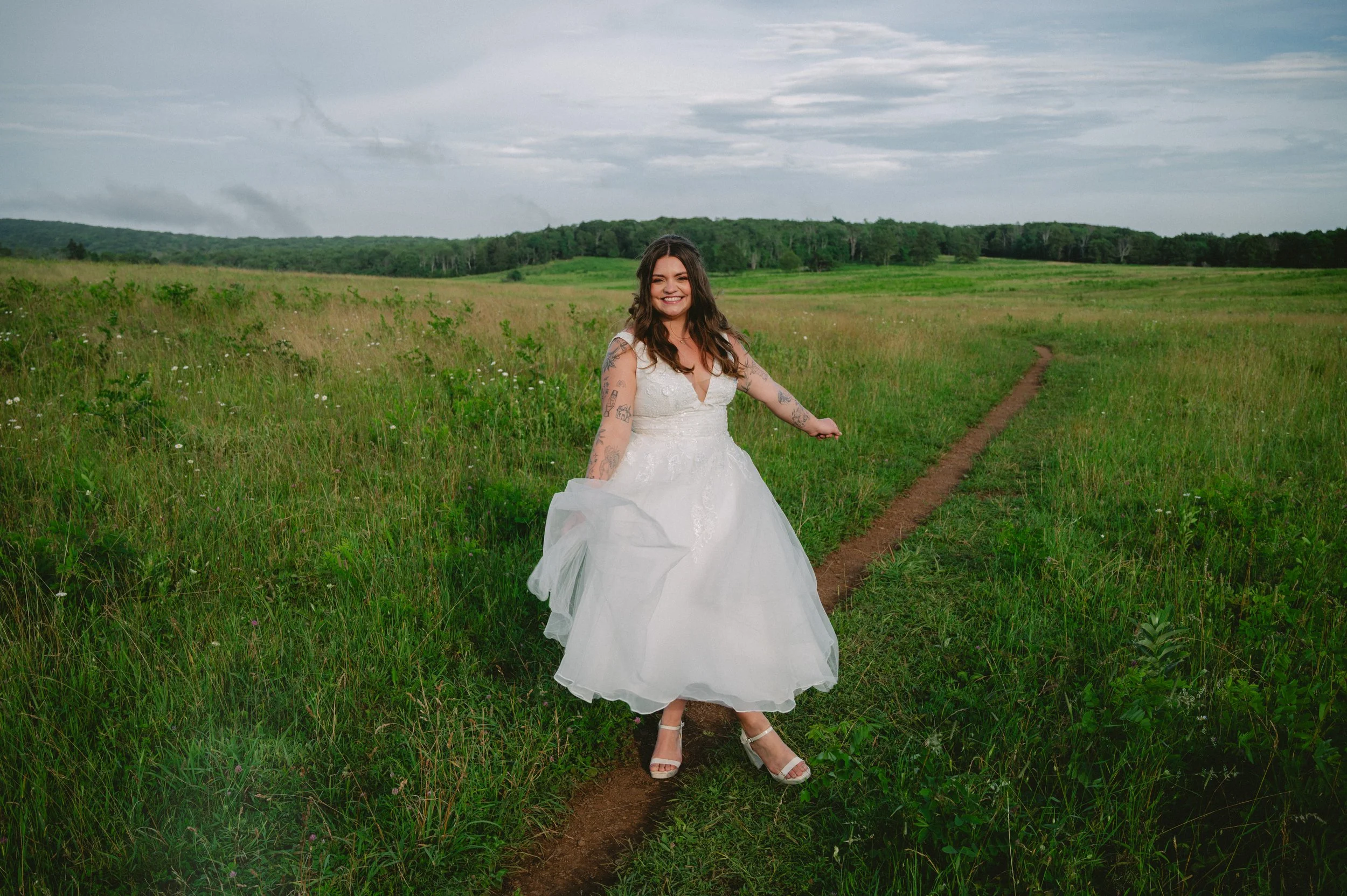 A woman in a white wedding dress walking on a narrow dirt trail through a green field with a cloudy sky in the background.