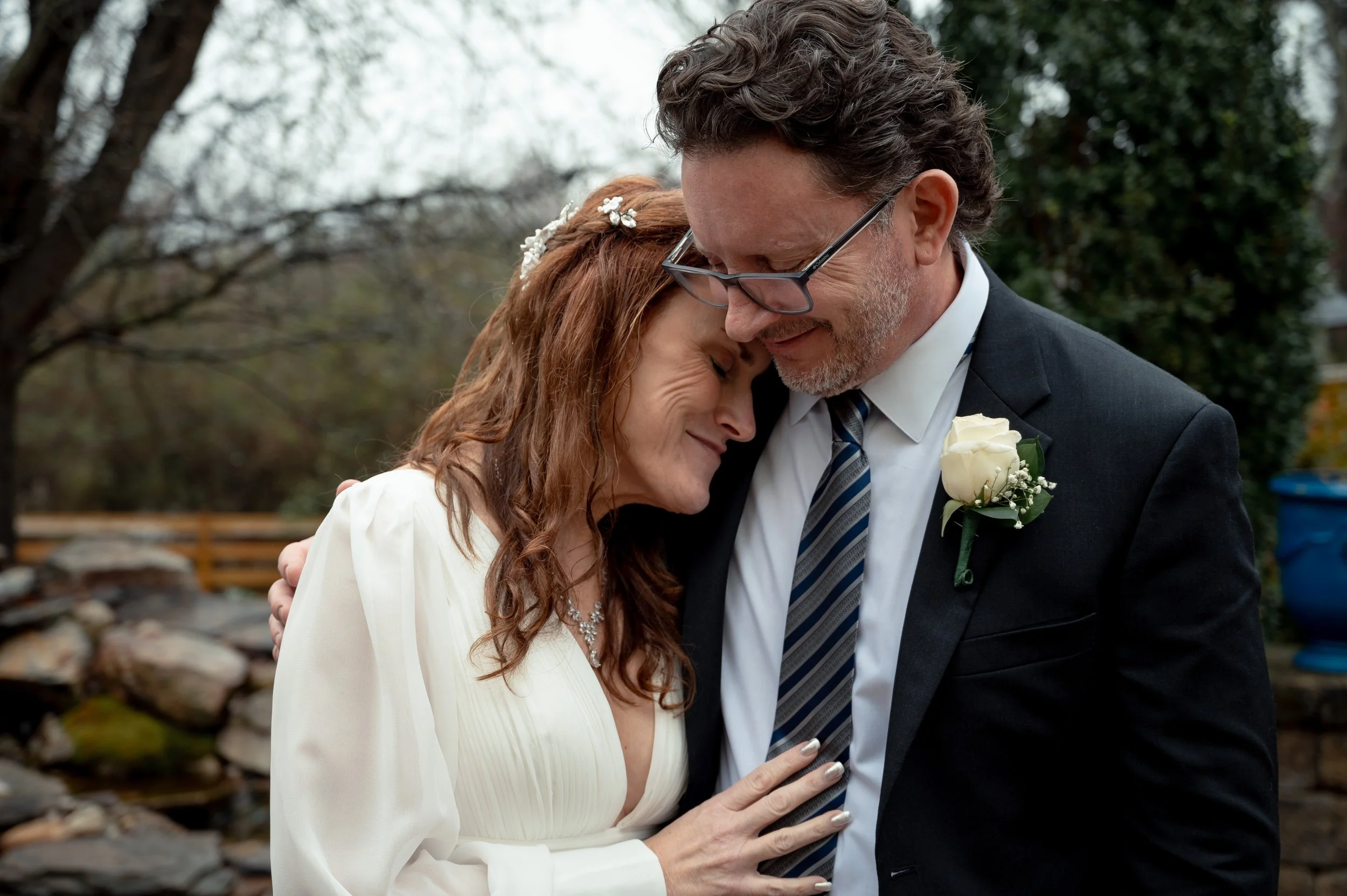 A middle-aged woman and an older man share a tender moment outdoors, with their foreheads touching and eyes closed, in a peaceful natural setting.