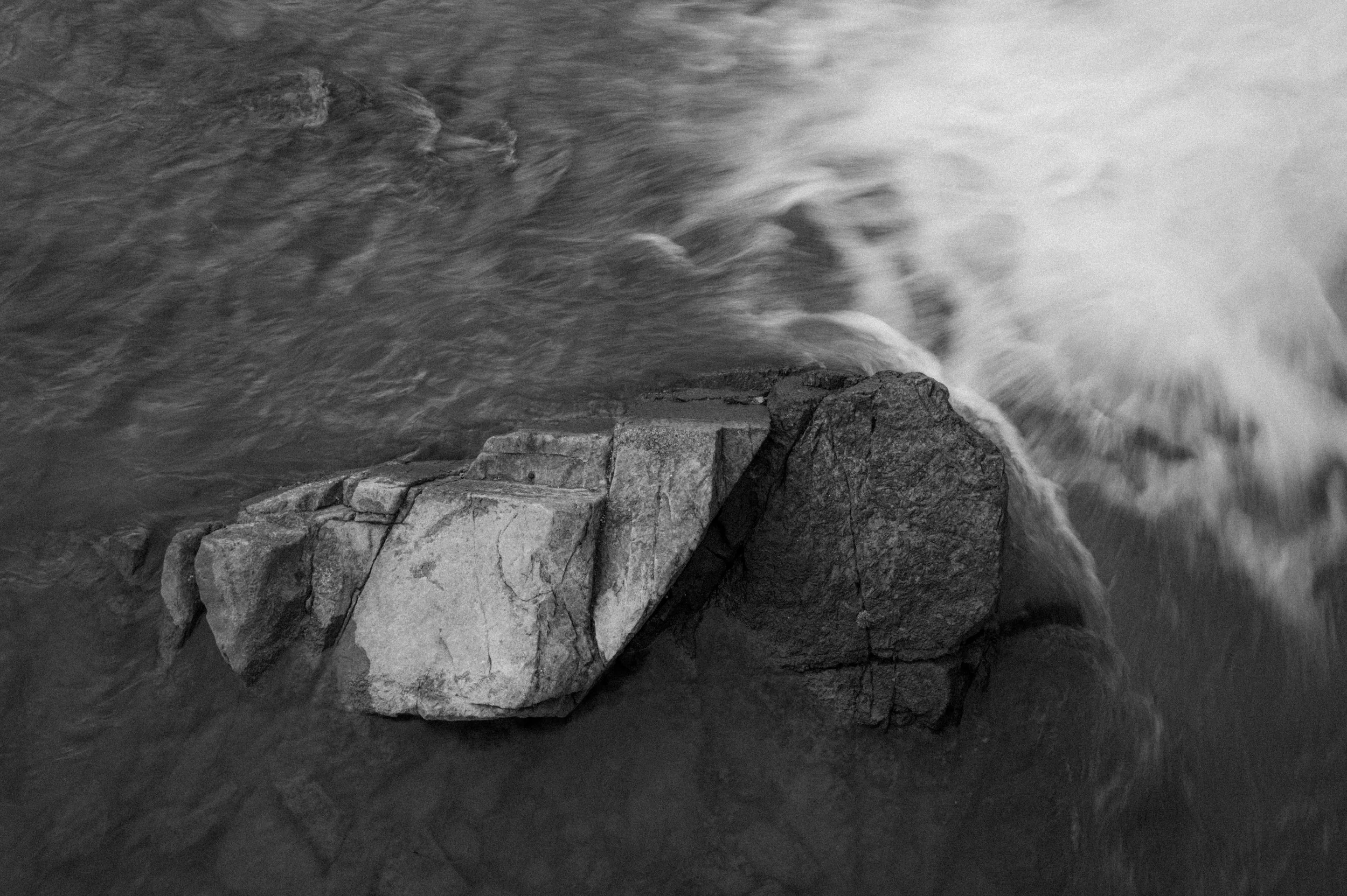 Black and white photograph of a large rock in a rushing stream or river.