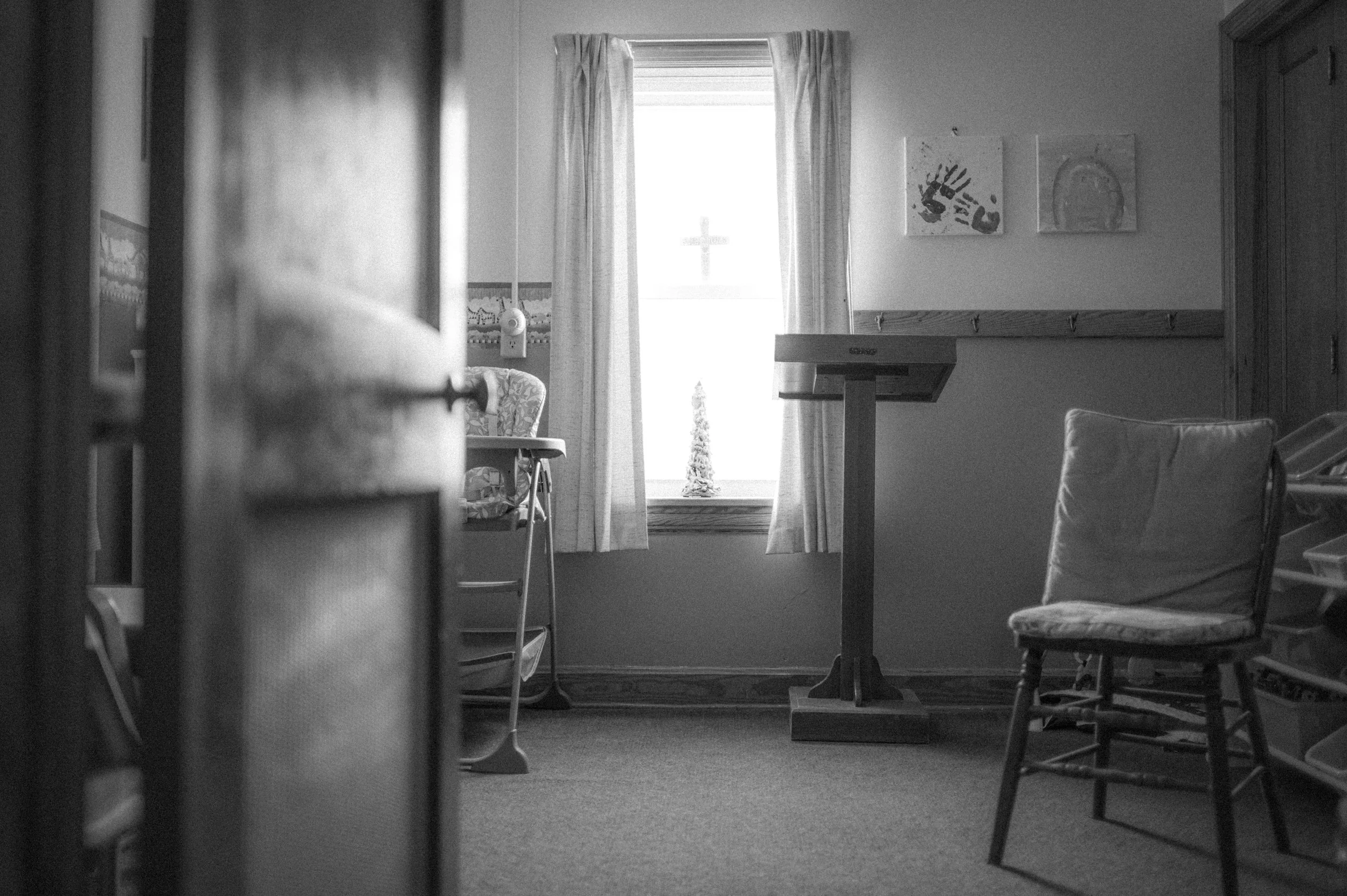 A black and white photo of a hospital or care room. Visible are a window with curtain, a rocking chair, a small table, and a wall with children's drawings, a coat rack, and medical equipment.