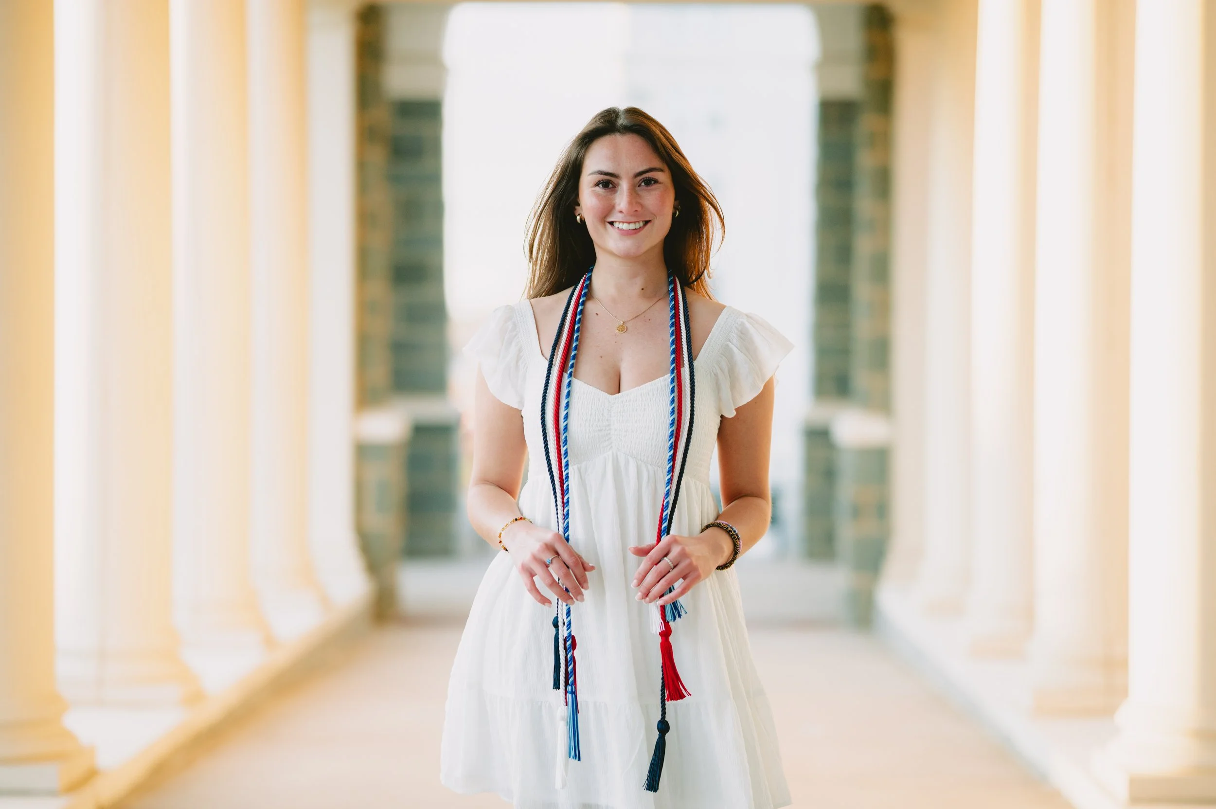 A young woman in a white dress smiling, standing in a bright hallway with columns, wearing a graduation stole around her neck.