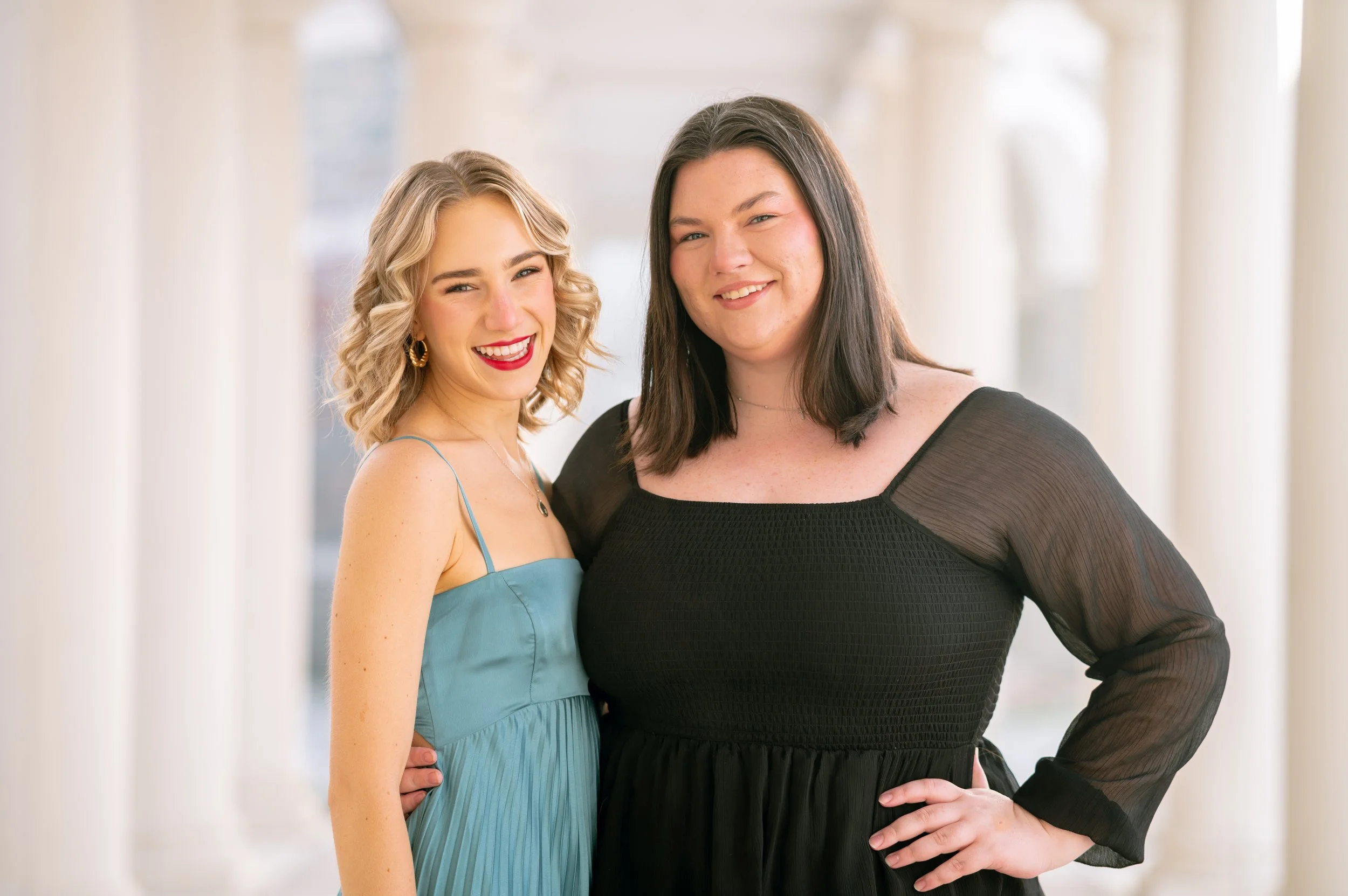 Two women standing happily together indoors, one with curly blonde hair in a blue dress, the other with straight dark hair in a black dress with sheer sleeves.