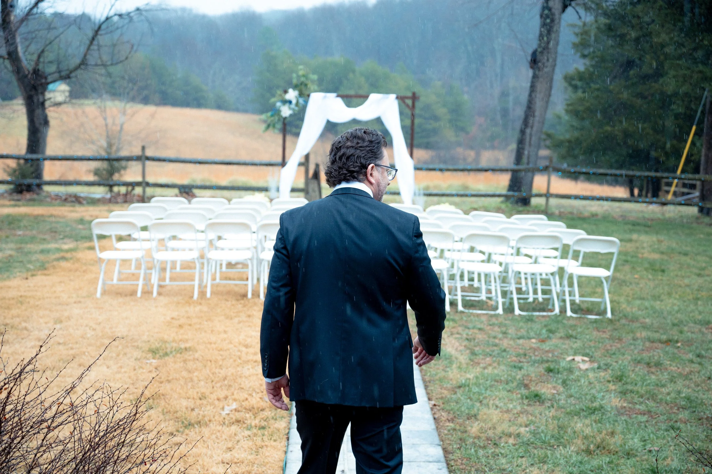 A man in a black suit and glasses walking toward a white wedding arch with white chairs arranged in rows outdoors on a rainy day