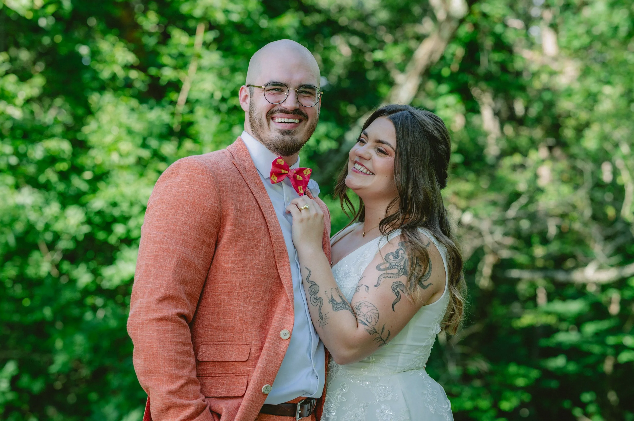 A smiling couple in wedding attire standing outdoors with green trees in the background.