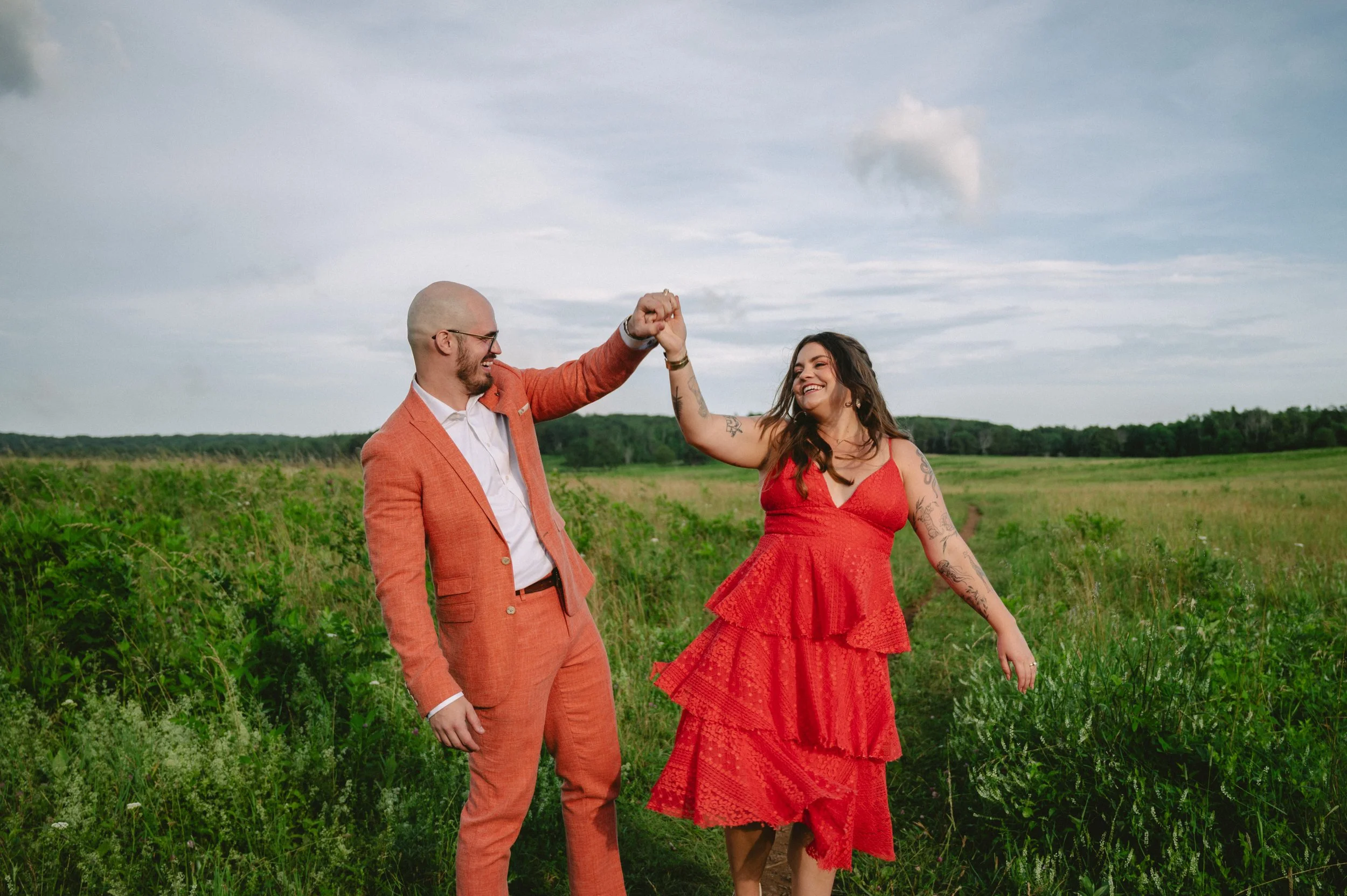 A man and woman in a green field, smiling and holding hands, with the man wearing an orange suit and the woman in a red dress.