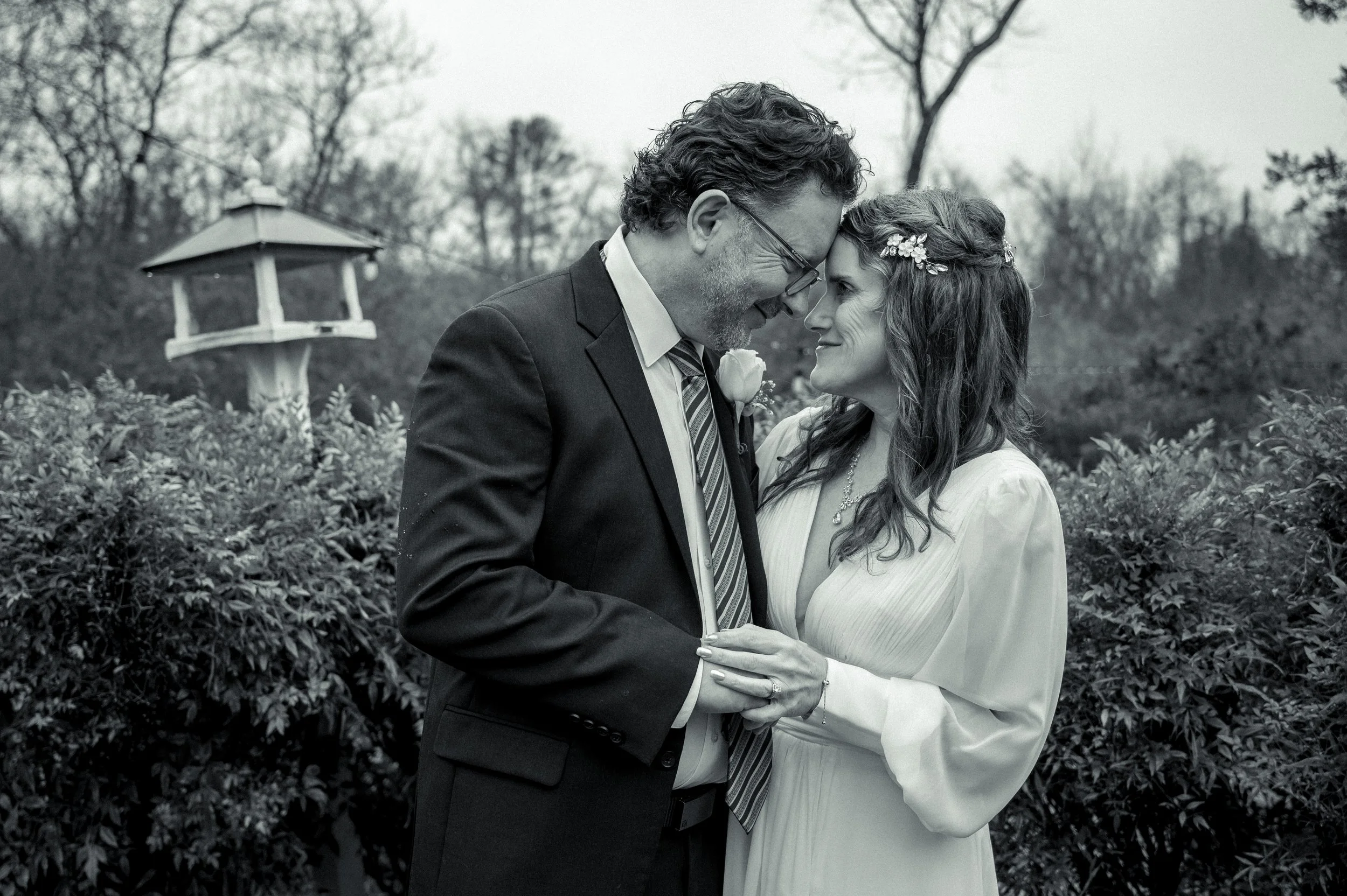 A black-and-white photo of a couple in wedding attire sharing a tender moment outdoors, with a garden and a birdhouse in the background.