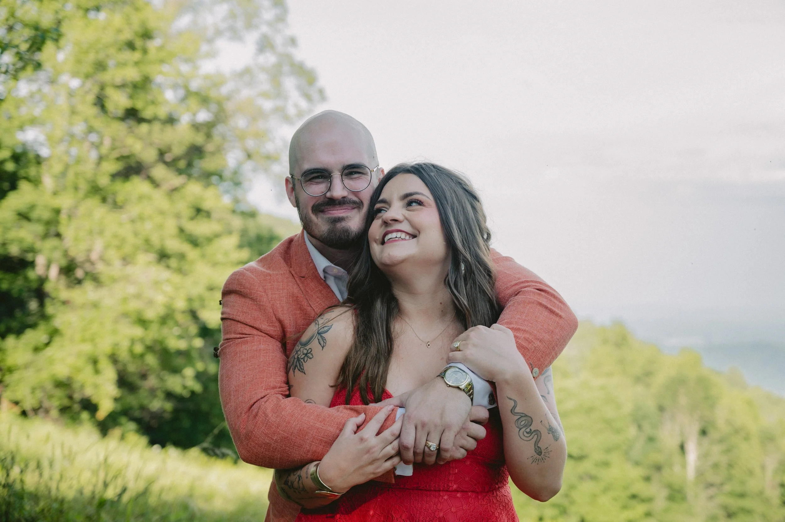 A couple embracing outdoors, smiling, with trees and a cloudy sky in the background. The man has a beard, glasses, and is wearing a peach blazer. The woman has long dark hair, tattoos on her arms, and is wearing a red dress. They are holding each oth