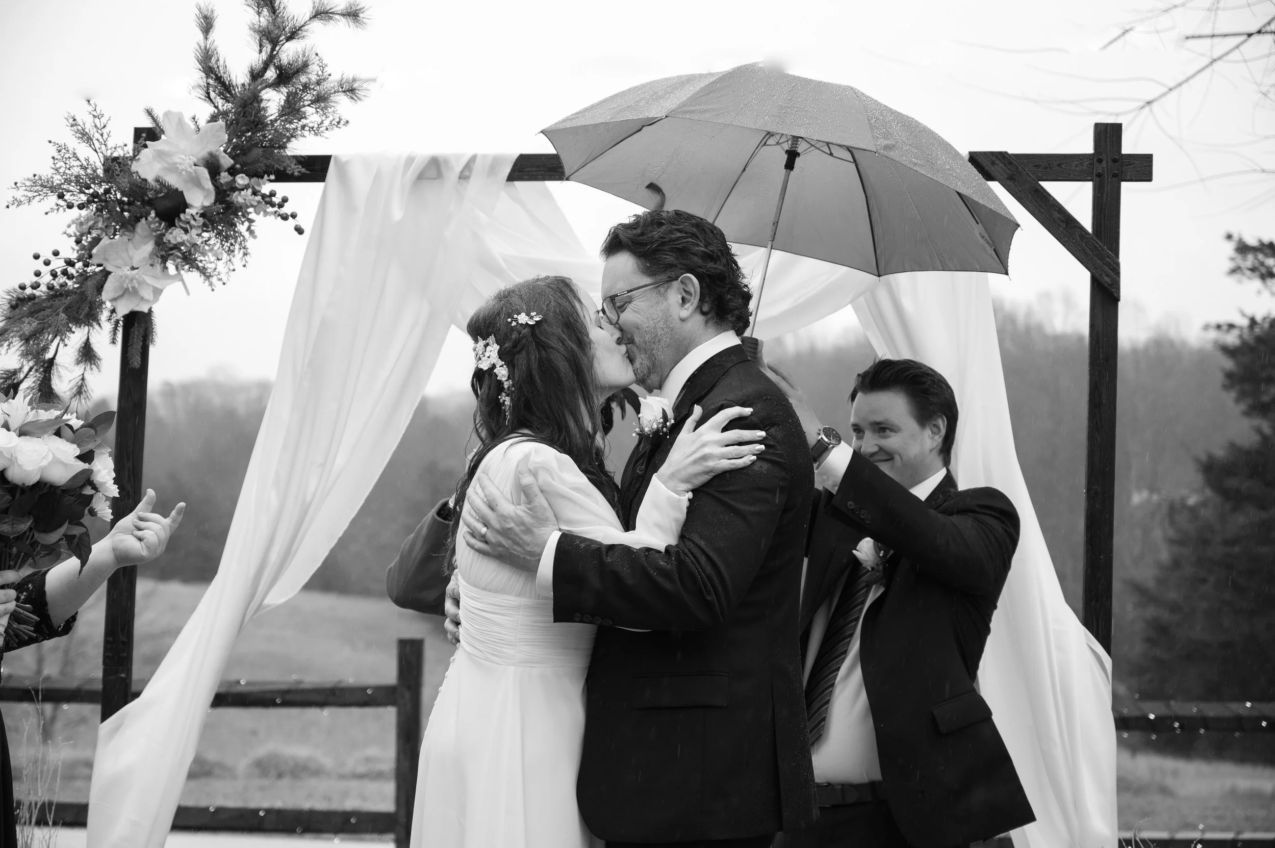 A black and white photo of a wedding ceremony where a couple is kissing under an umbrella, with two men in suits, one holding the umbrella, standing on a decorated outdoor wooden arch.