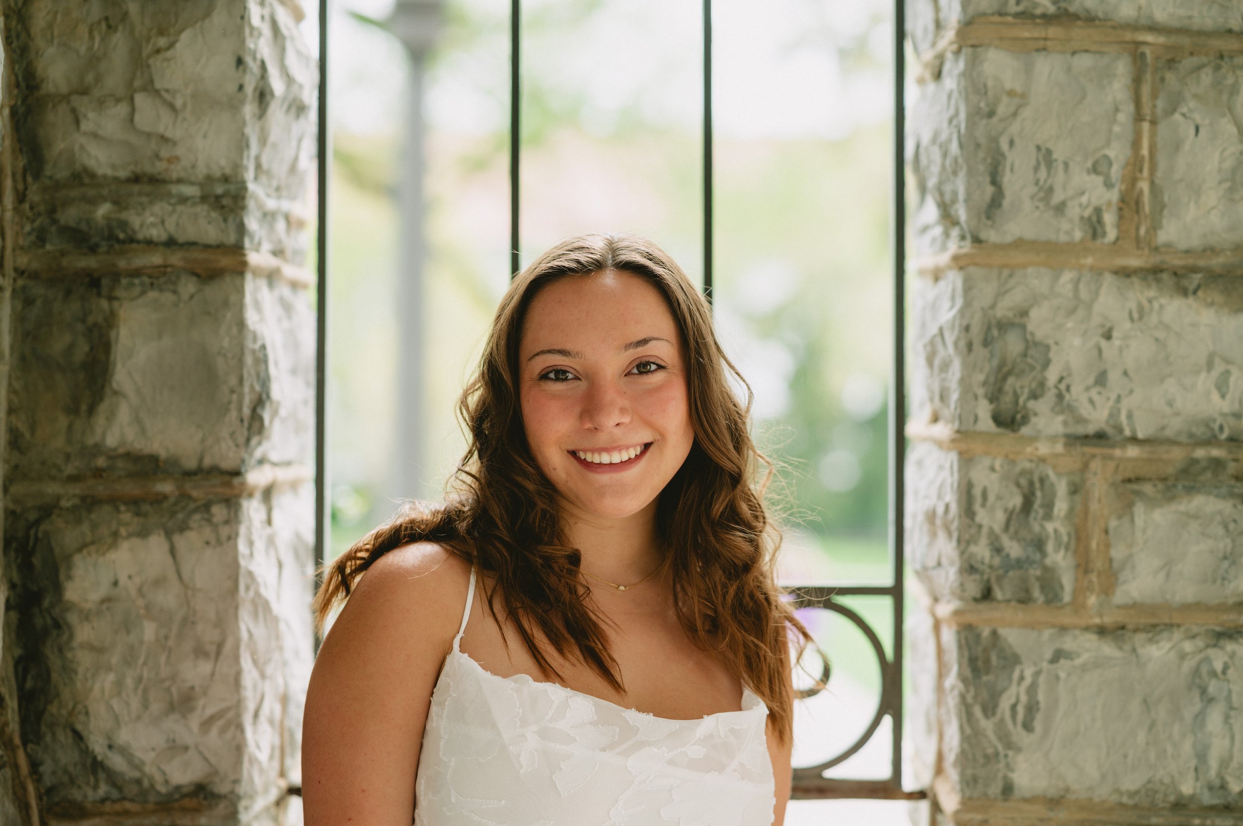 A young woman with brown wavy hair and a white dress smiling, standing in front of a stone wall with a glass window behind her.