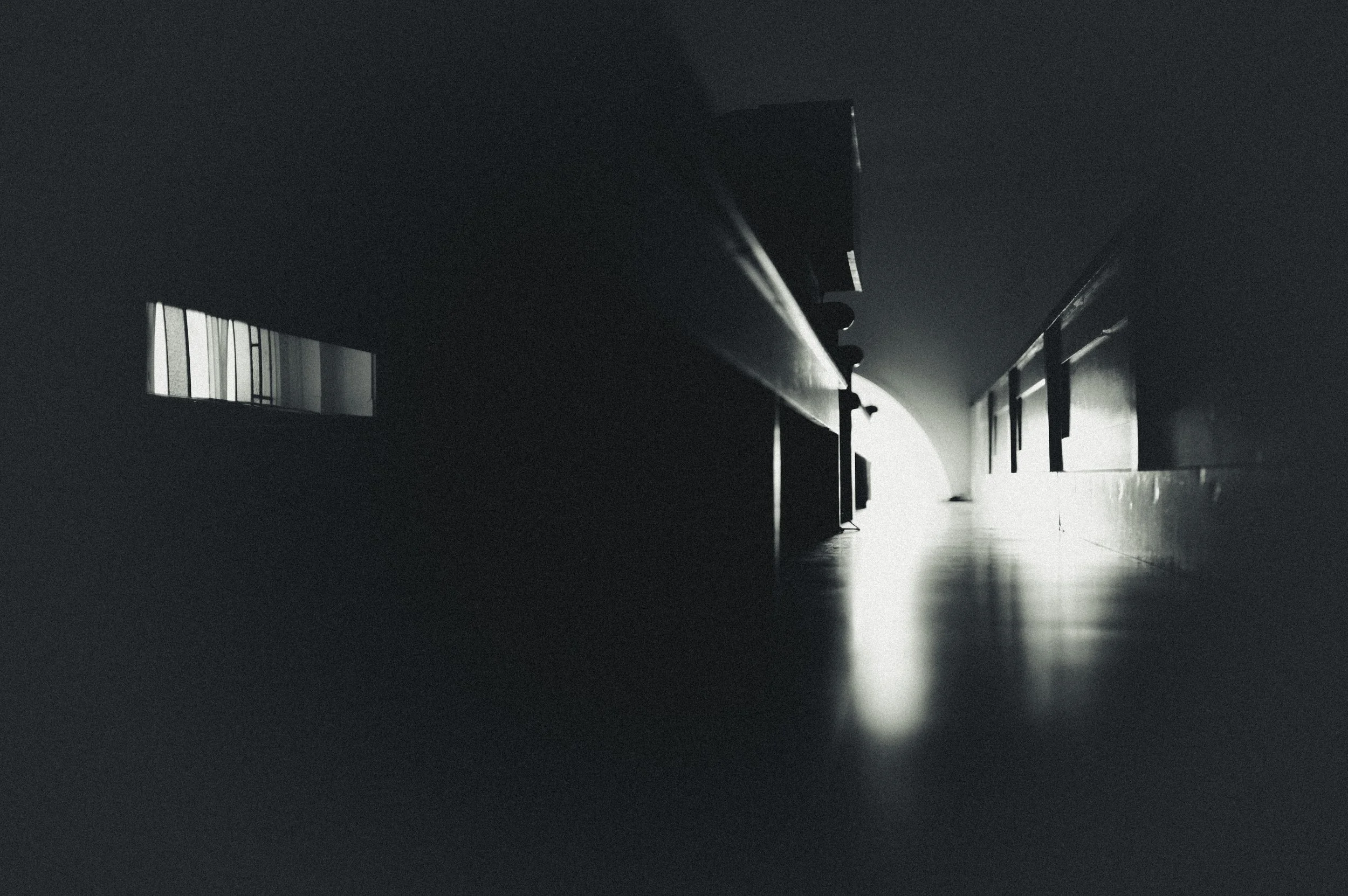 A dark, low-angle view of a corridor with bright light at the end, showing windows on the left side and picture frames on the right wall.