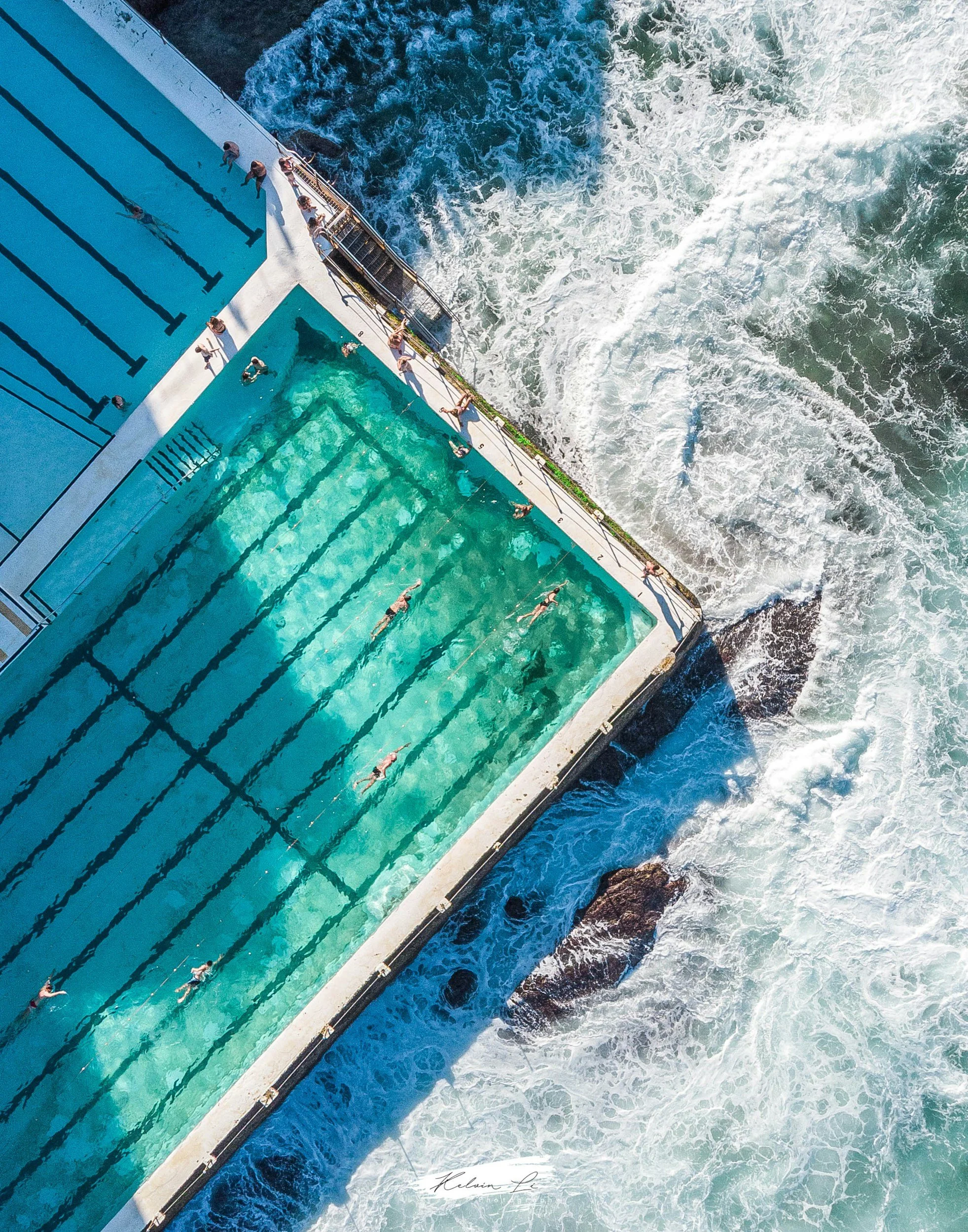 Aerial view of a swimming pool with swimmers, adjacent to an ocean with waves crashing against the pool edge.